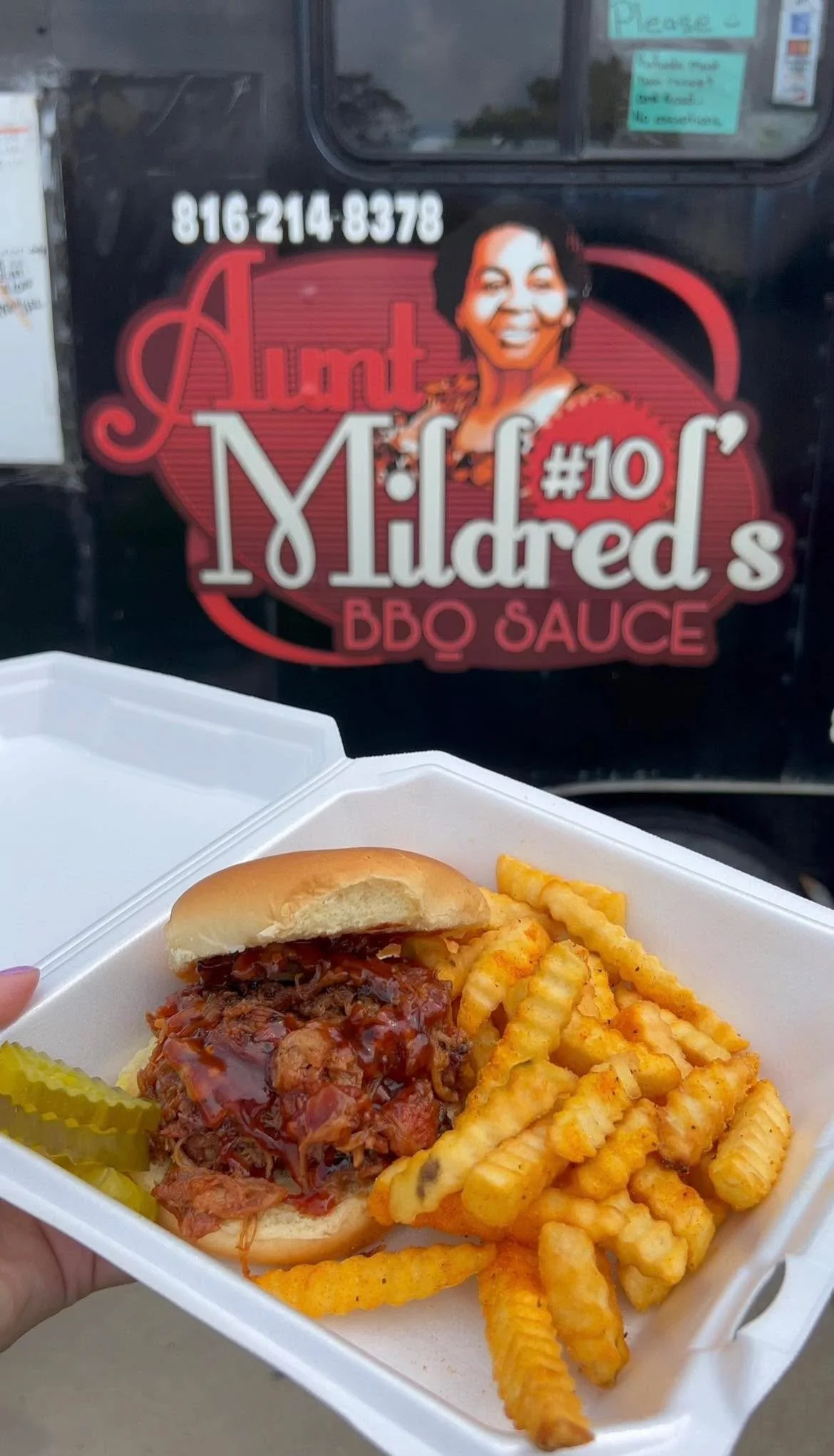 A takeout container holding a barbecue sandwich with coleslaw in a bun, crinkle-cut French fries seasoned with spices, and pickle slices. In the background, a food truck displays a sign for Aunt Mildred's BBQ Sauce with a picture of a smiling woman.