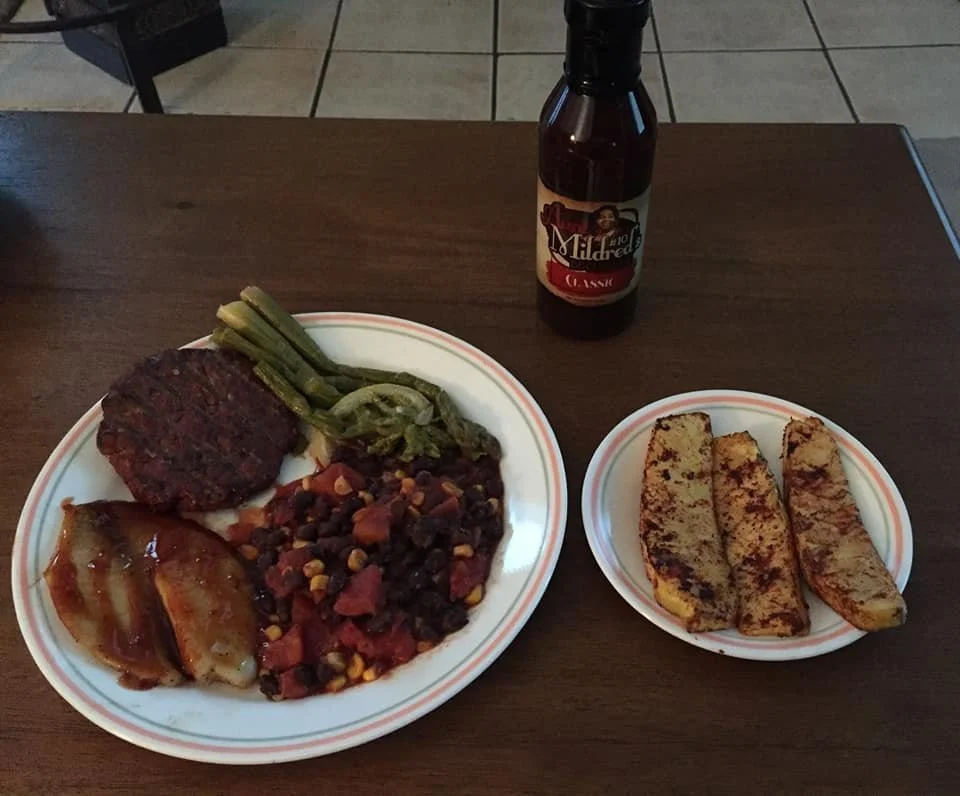 A plate of food with grilled hamburger patty, green beans, baked chicken with sauce, and a bean and vegetable chili, with a side of three pieces of French toast, and a bottle of maple syrup on a wooden table.