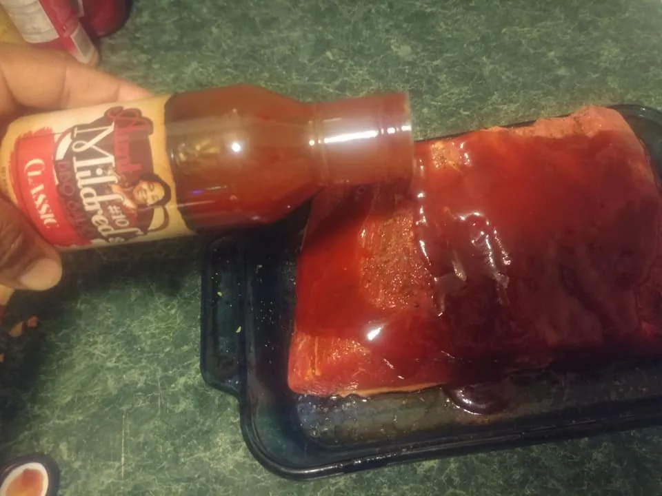 A package of pork ribs being coated with barbecue sauce in a black roasting pan.