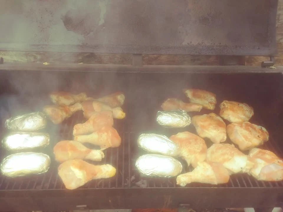 Chicken drumsticks and foil-wrapped potatoes cooking on a grill.