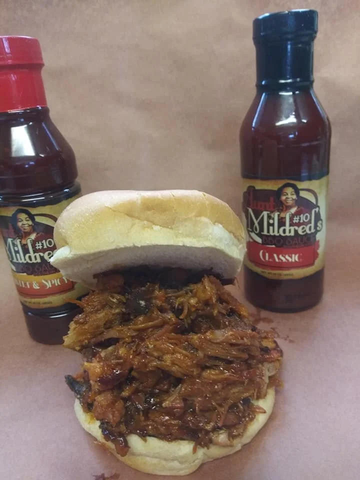 Barbecue pork sandwich in a bun with Mildred’s barbecue sauces in bottles in the background.
