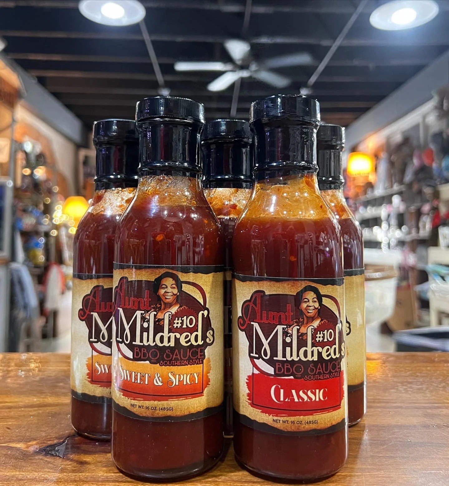 Six bottles of Aunt Mildred's BBQ sauce, with three labeled 'Sweet & Spicy' and three labeled 'Classic,' arranged on a wooden surface with a blurred store interior background.