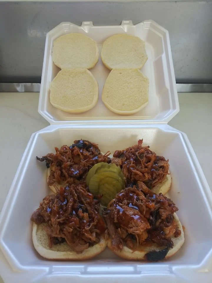 Four sandwich buns topped with shredded barbecue pulled pork and pickles, served in a white foam to-go container.