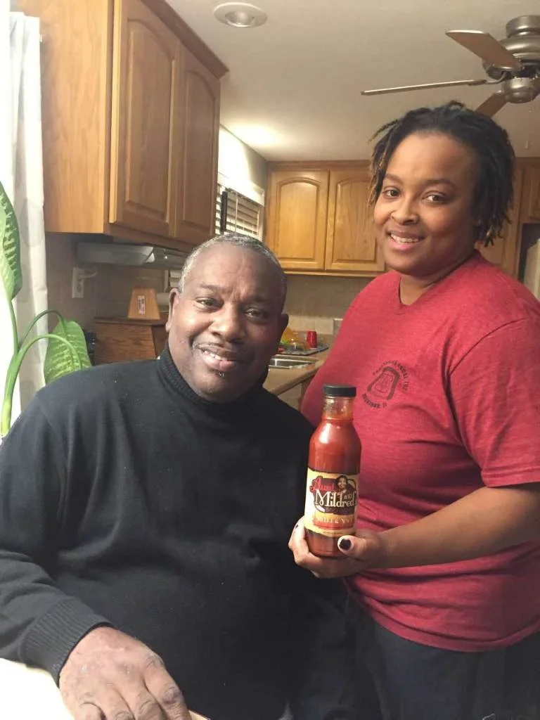 A man and a woman posing together in a kitchen, with the woman holding a bottle of hot sauce.