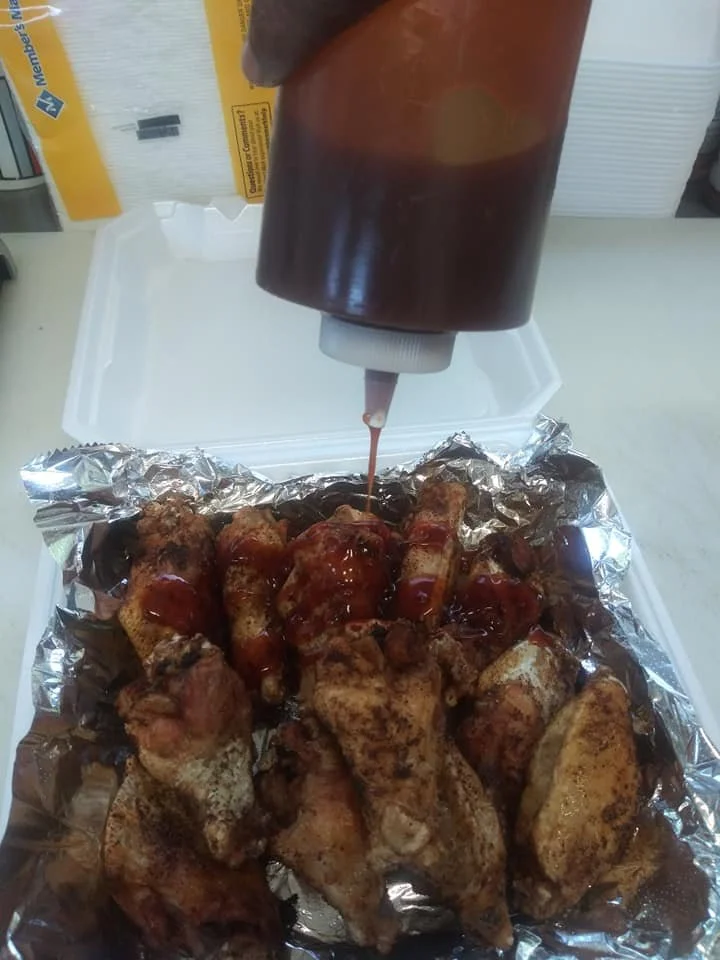 Fried chicken drumsticks with barbecue sauce being drizzled on top, placed on aluminum foil inside a white takeout container.