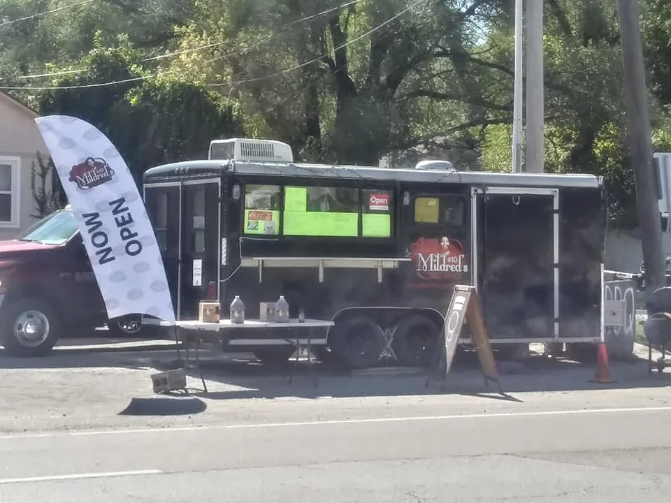 A black food truck named 'Milfred's' parked on the side of the street, with a white flag that says 'NOW OPEN' in front of it. There are orange cones, a sandwich board, and bottles on a small table by the truck. The background has trees and power line