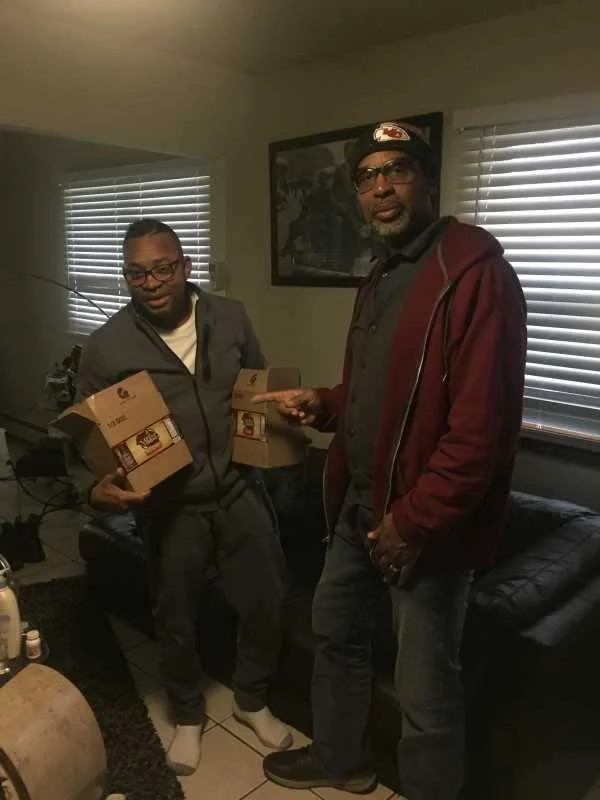 Two men standing in a living room holding boxes of food and an envelope, with windows, blinds, and a picture on the wall behind them.