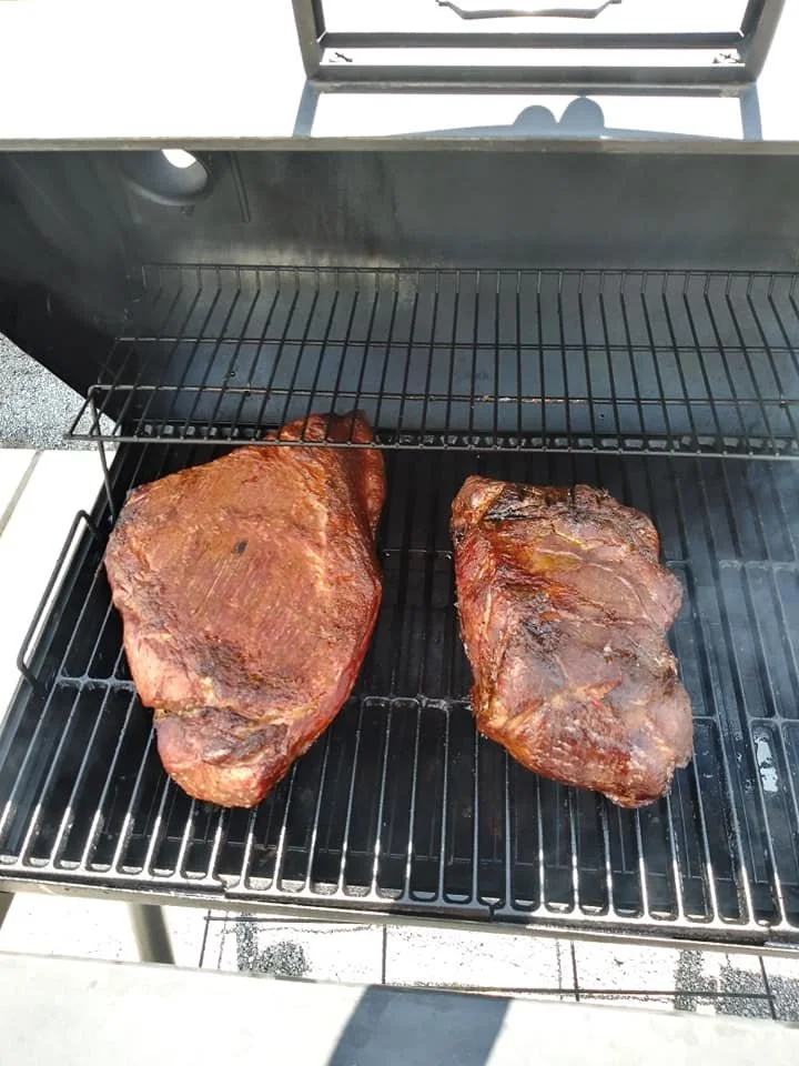 Two large seasoned beef briskets cooking on a black outdoor grill over a heated element.
