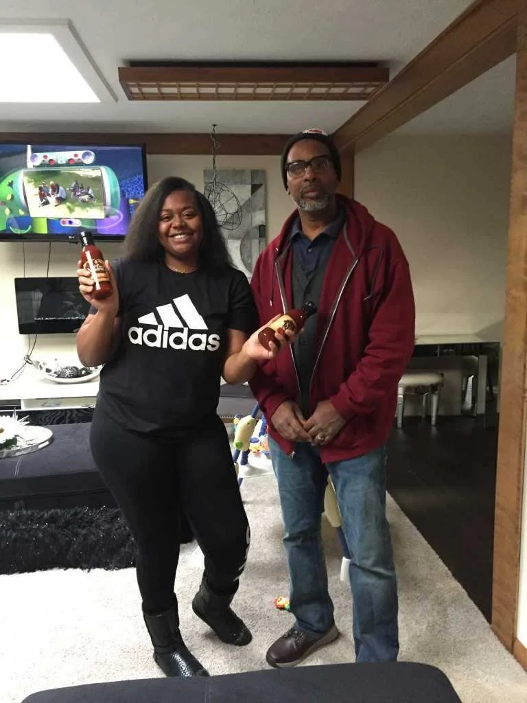 A woman and a man stand indoors holding bottles of sauce, smiling and posing for the photo. The woman is wearing a black Adidas t-shirt, black pants, and boots. The man is wearing glasses, a beanie, a red jacket over a dark shirt, and jeans. There is
