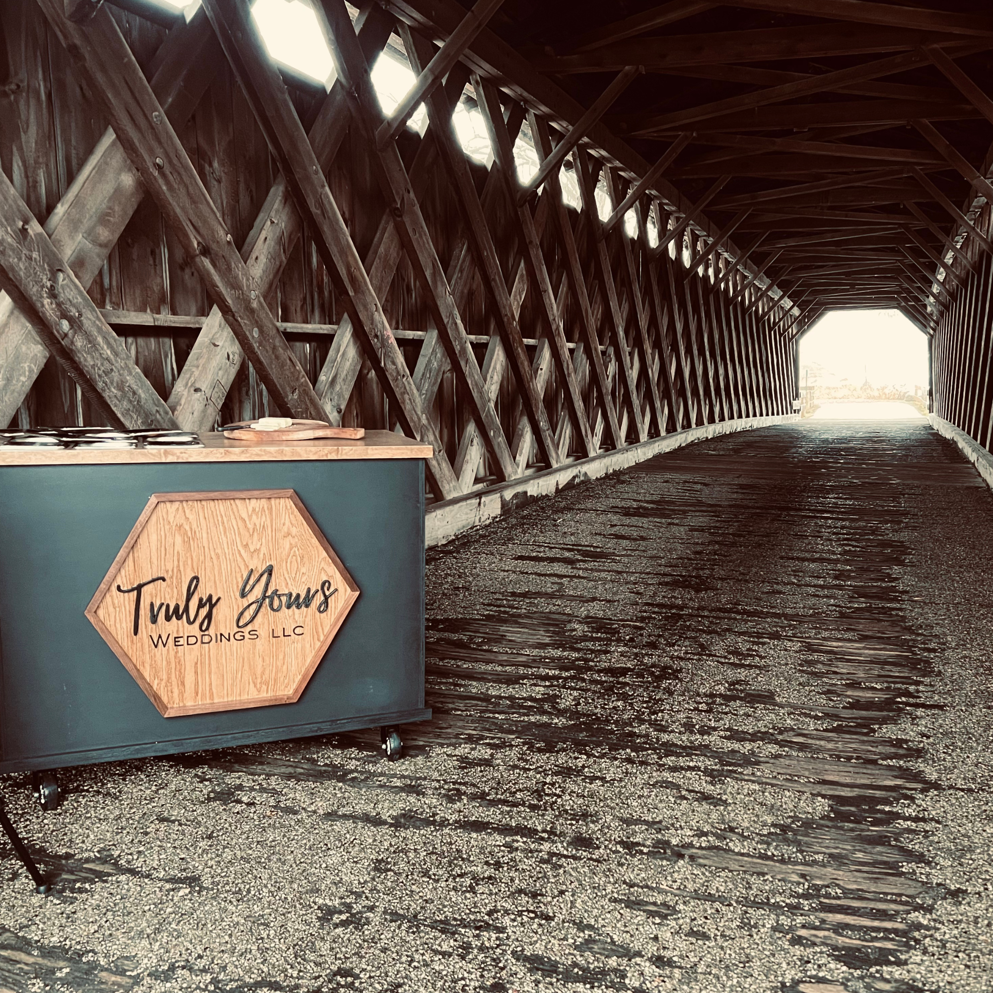 A portable event sign on wheels inside a rustic wooden tunnel. The sign reads 'Truly Young Weddings LLC' with a hexagonal wooden background and black cursive letters. The tunnel has a wooden arched ceiling and walls with diagonal support beams, and sunlight is seen at the far end of the tunnel.