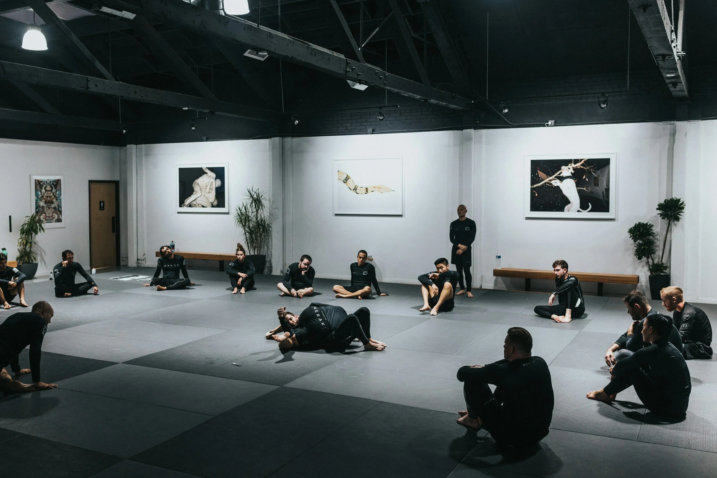 People practicing Brazilian Jiu-Jitsu in an indoor gym, some sitting on the mat, and one demonstrating a ground fighting technique while others observe.