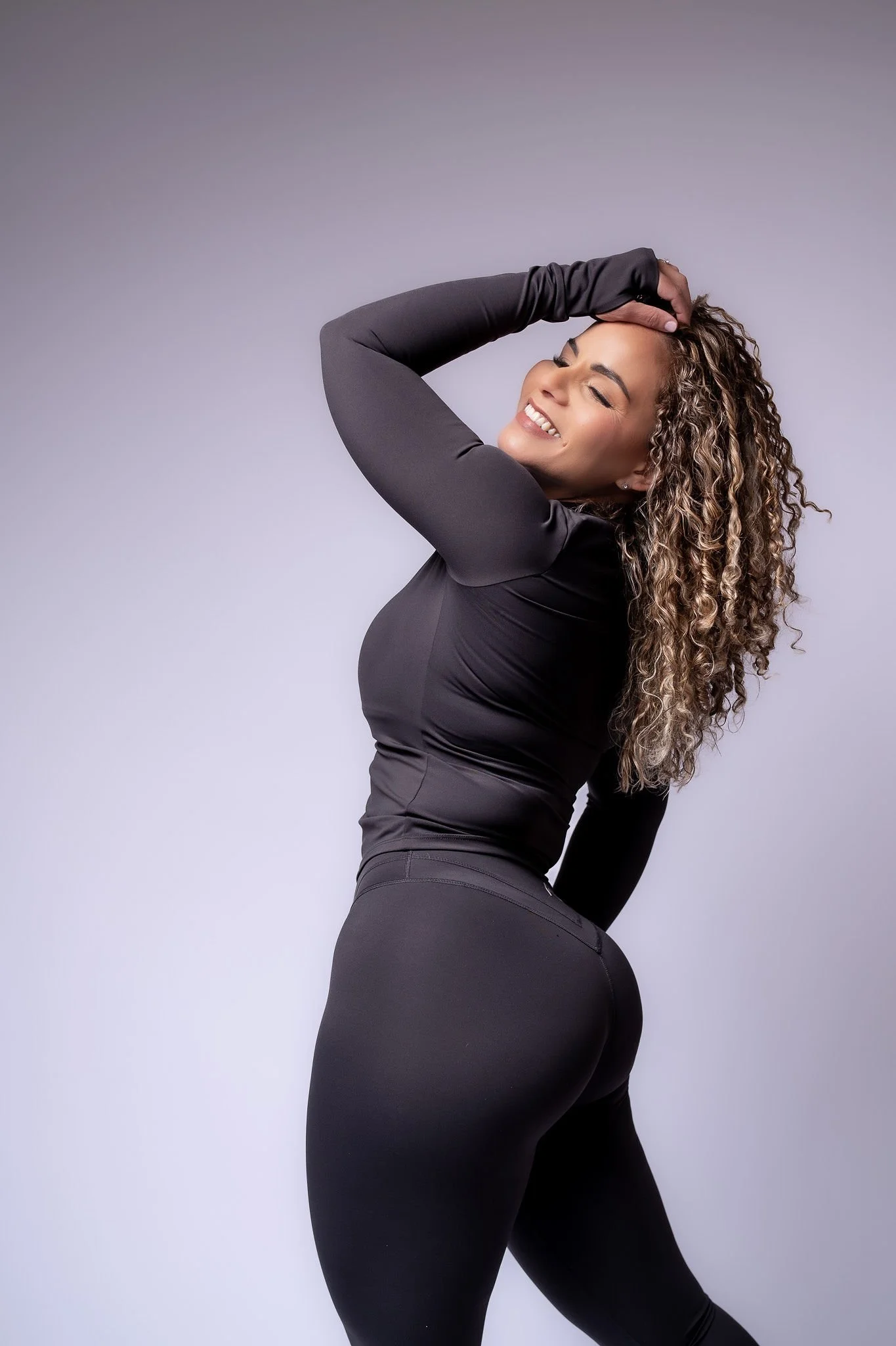 A woman with curly hair, smiling and posing with her hand on her head against a plain light background, wearing black athletic clothing.
