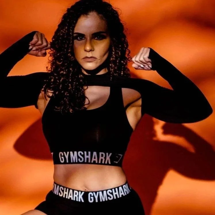 A woman with curly hair in a black sports bra and matching cropped leggings with Gymshark branding, flexing her biceps against an orange sunset background.