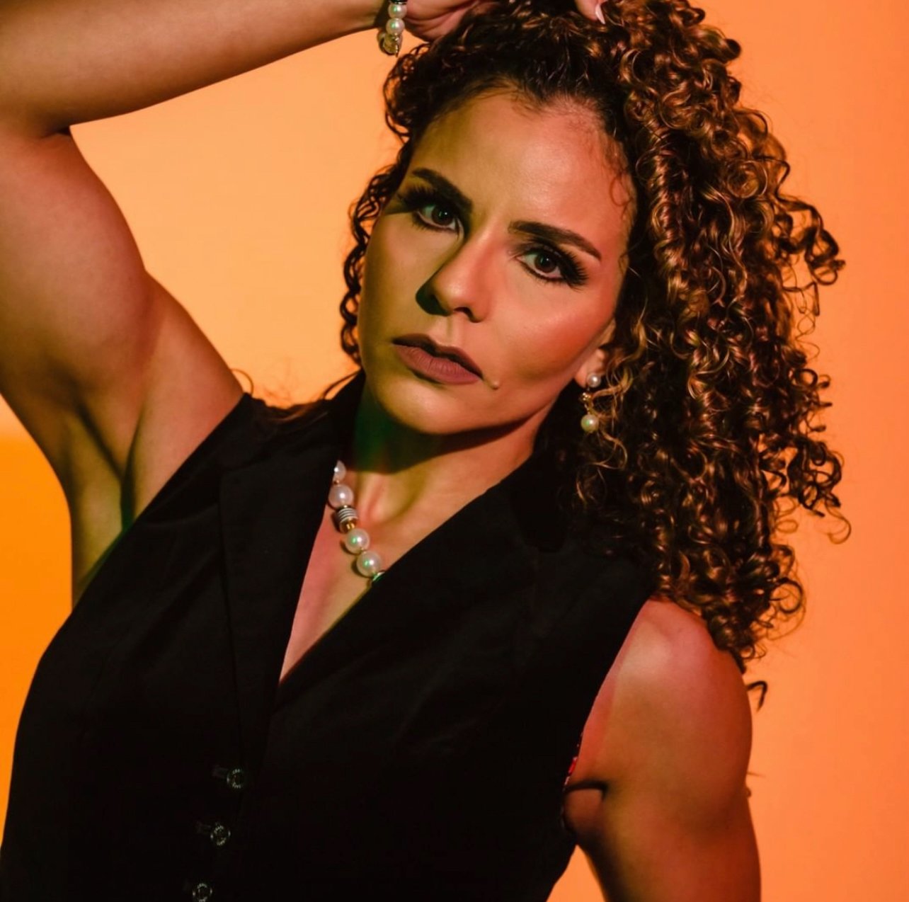 A woman with curly hair posing against a warm, colorful background. She is wearing a black sleeveless top, pearl earrings, and a pearl necklace. She has one arm raised, with her hand resting on her head.