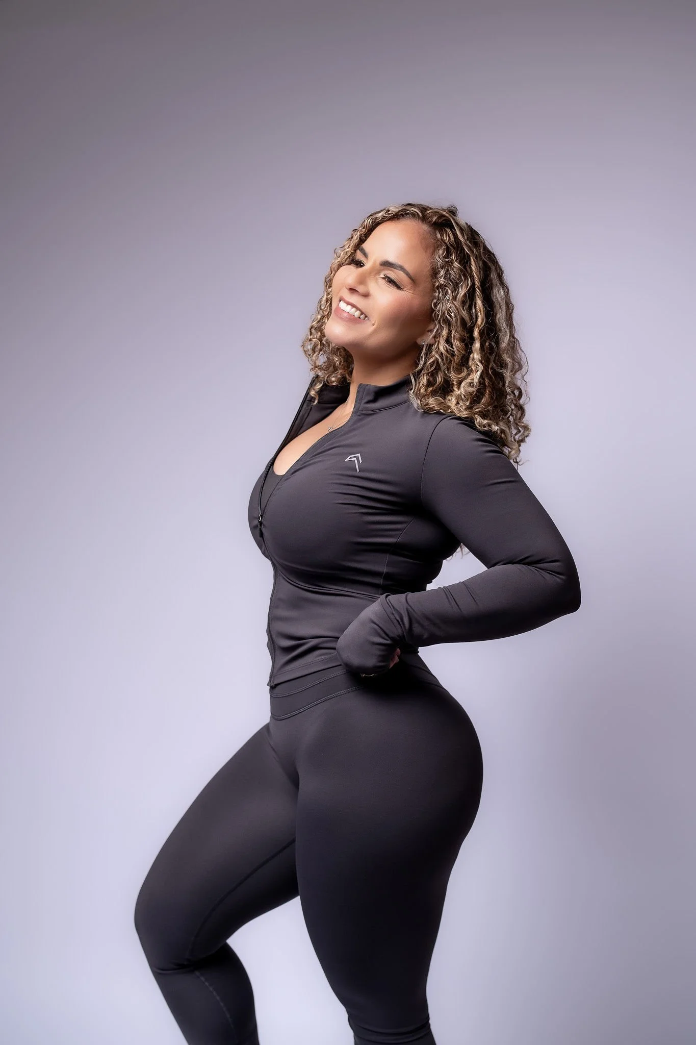 Woman with curly hair in black athletic wear smiling against plain background.