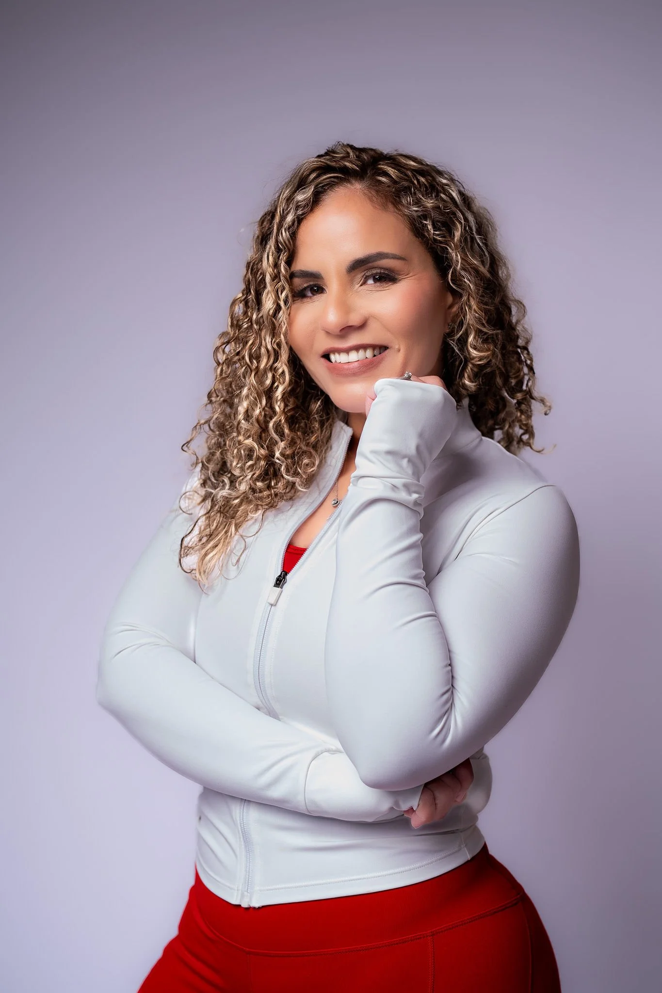 Judith smiling with curly hair wearing a white zip-up jacket and red pants, standing against a light gray background.