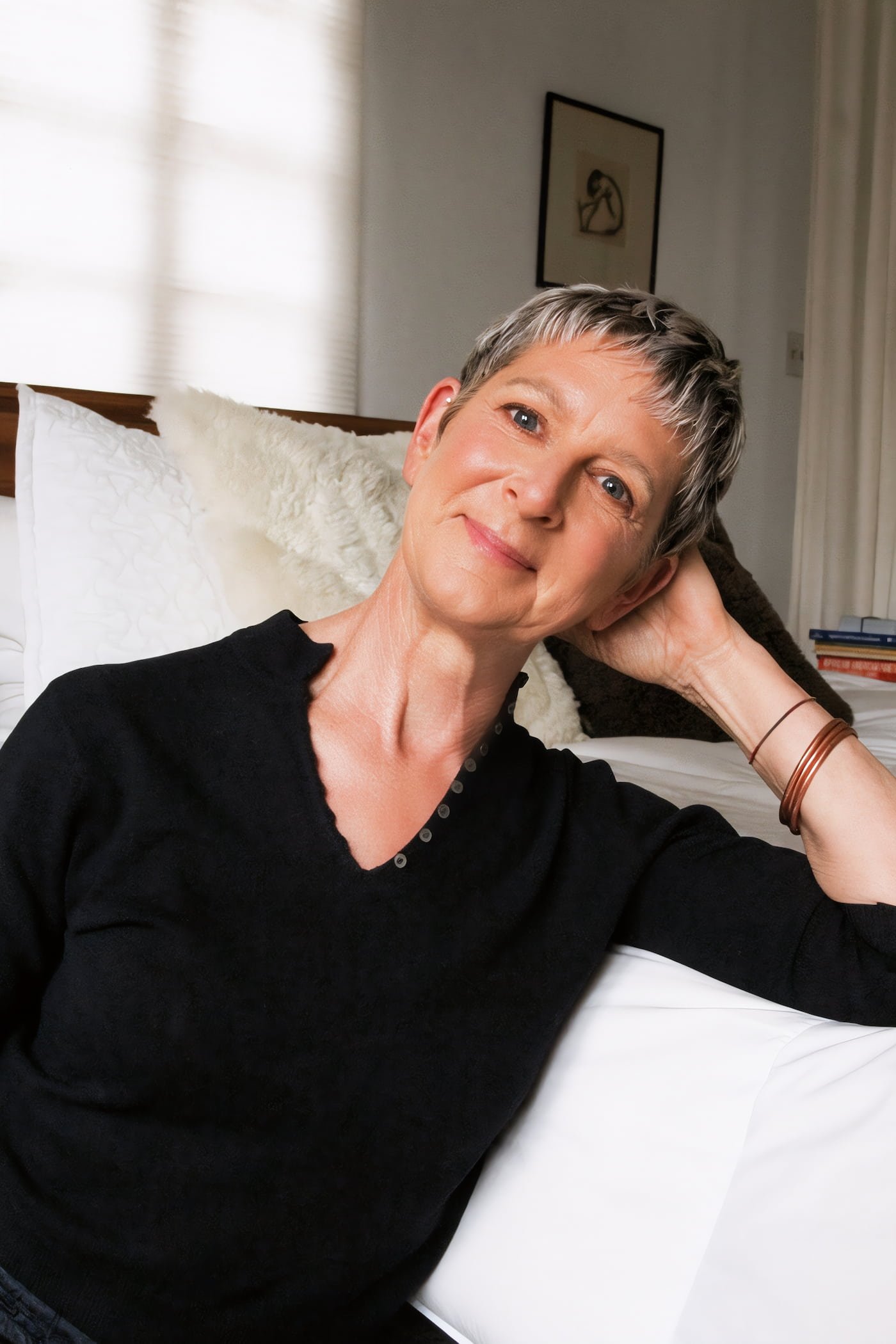 A woman with short gray hair sitting on a bed, wearing a black top, smiling, with her hand resting on her head in a cozy bedroom.