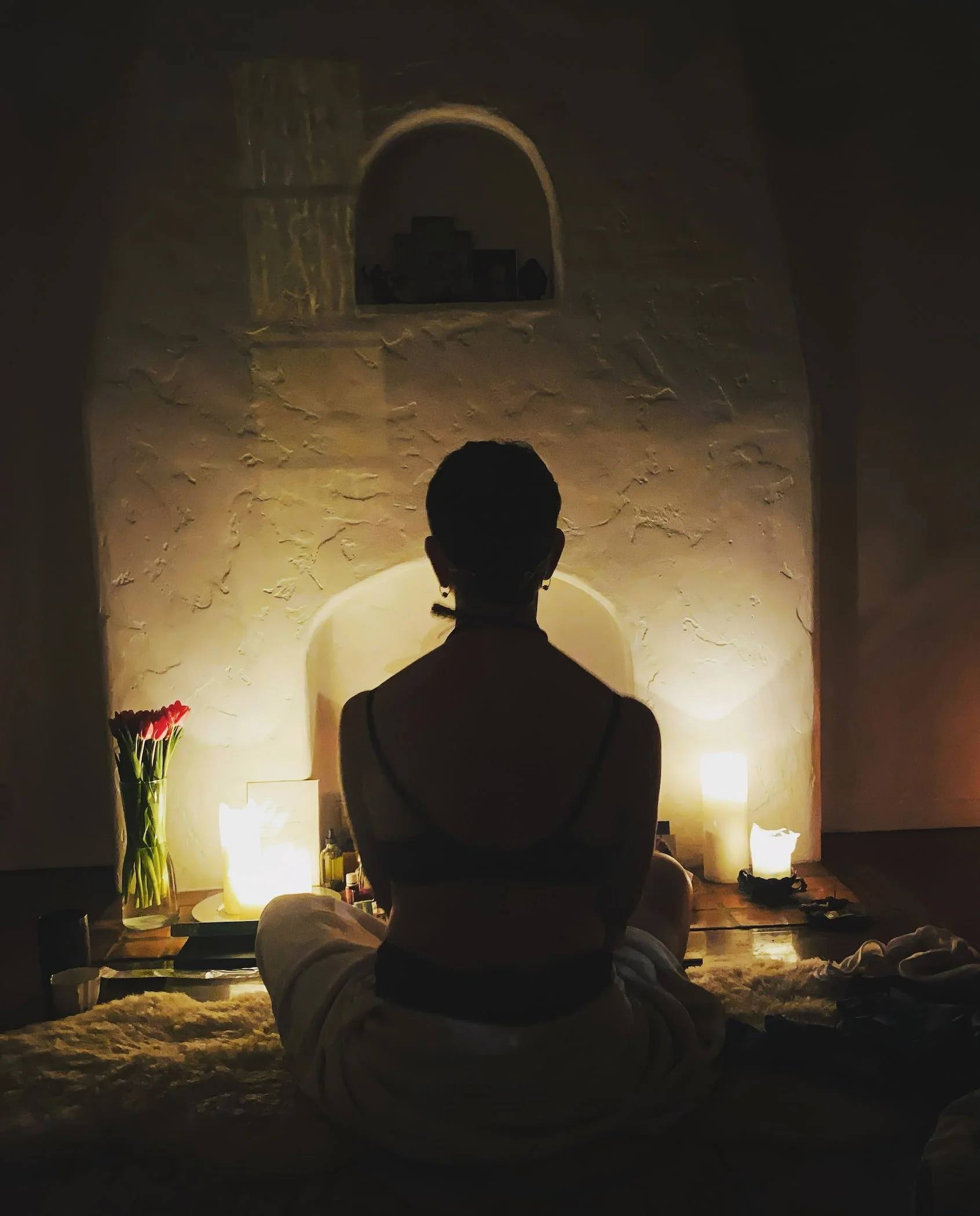 A person sitting cross-legged on the floor facing a lit wall with an arch-shaped window. The room is dimly lit with soft lights, some flowers on the left, and various objects on the right.