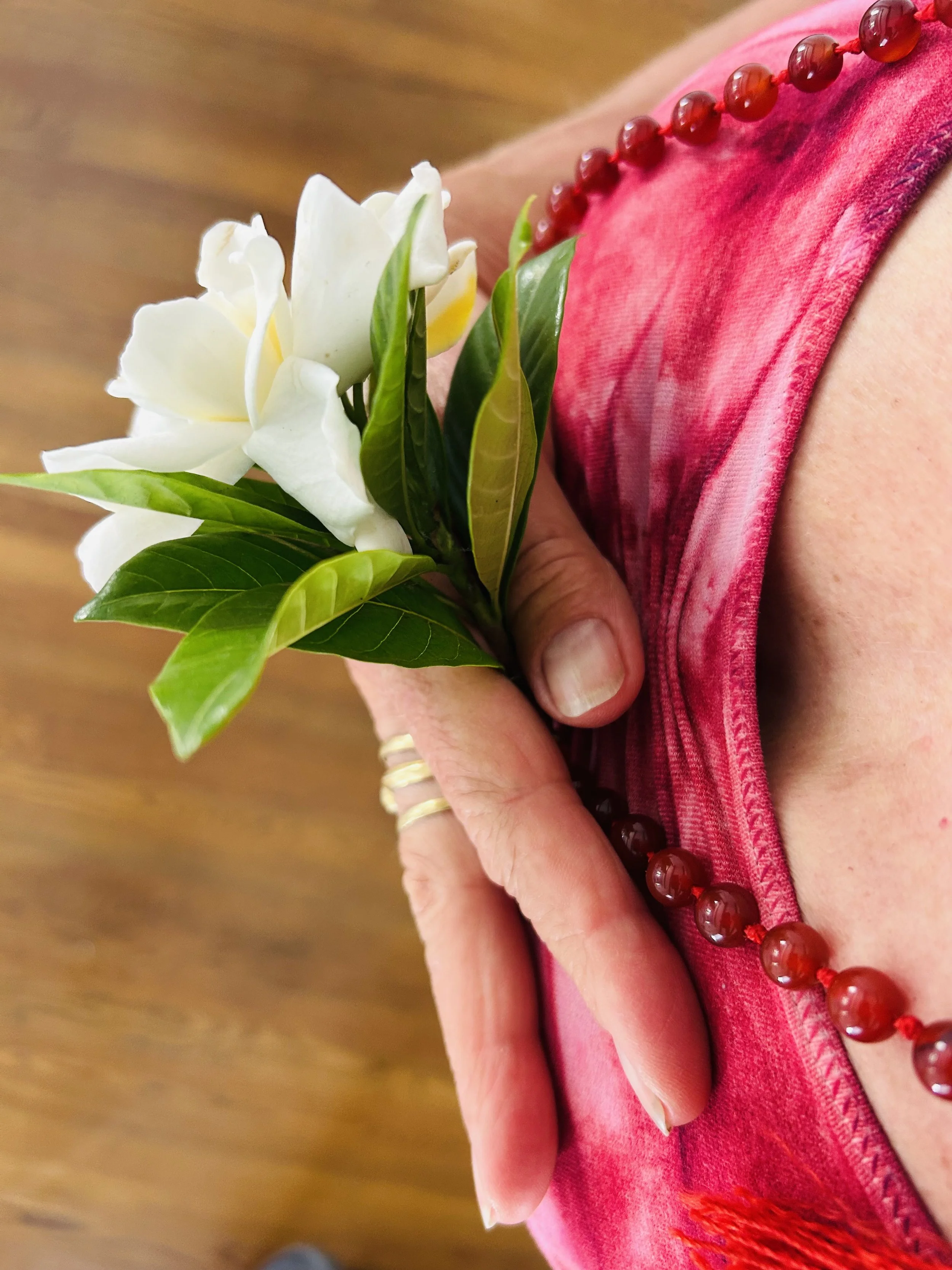 Close-up of a person's shoulder wearing a pink garment, with a white flower and green leaves tucked into the fabric, and a red beaded necklace.