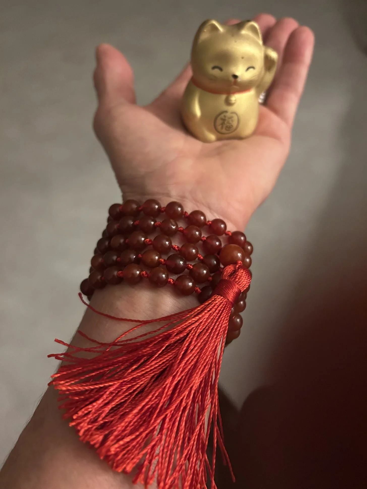 A hand with multiple red prayer beads and orange tassels holding a small gold cat-shaped figurine.