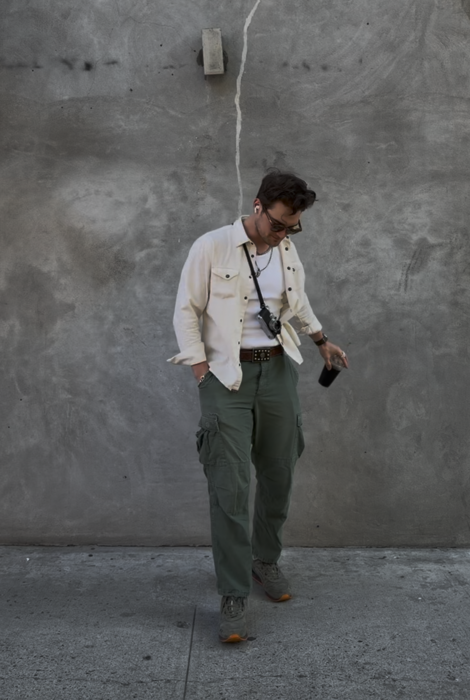 A man with glasses, a white jacket, a white t-shirt, green cargo pants, and sneakers stands in front of a gray concrete wall, holding a smartphone in his left hand and looking down, with a camera hanging around his neck.