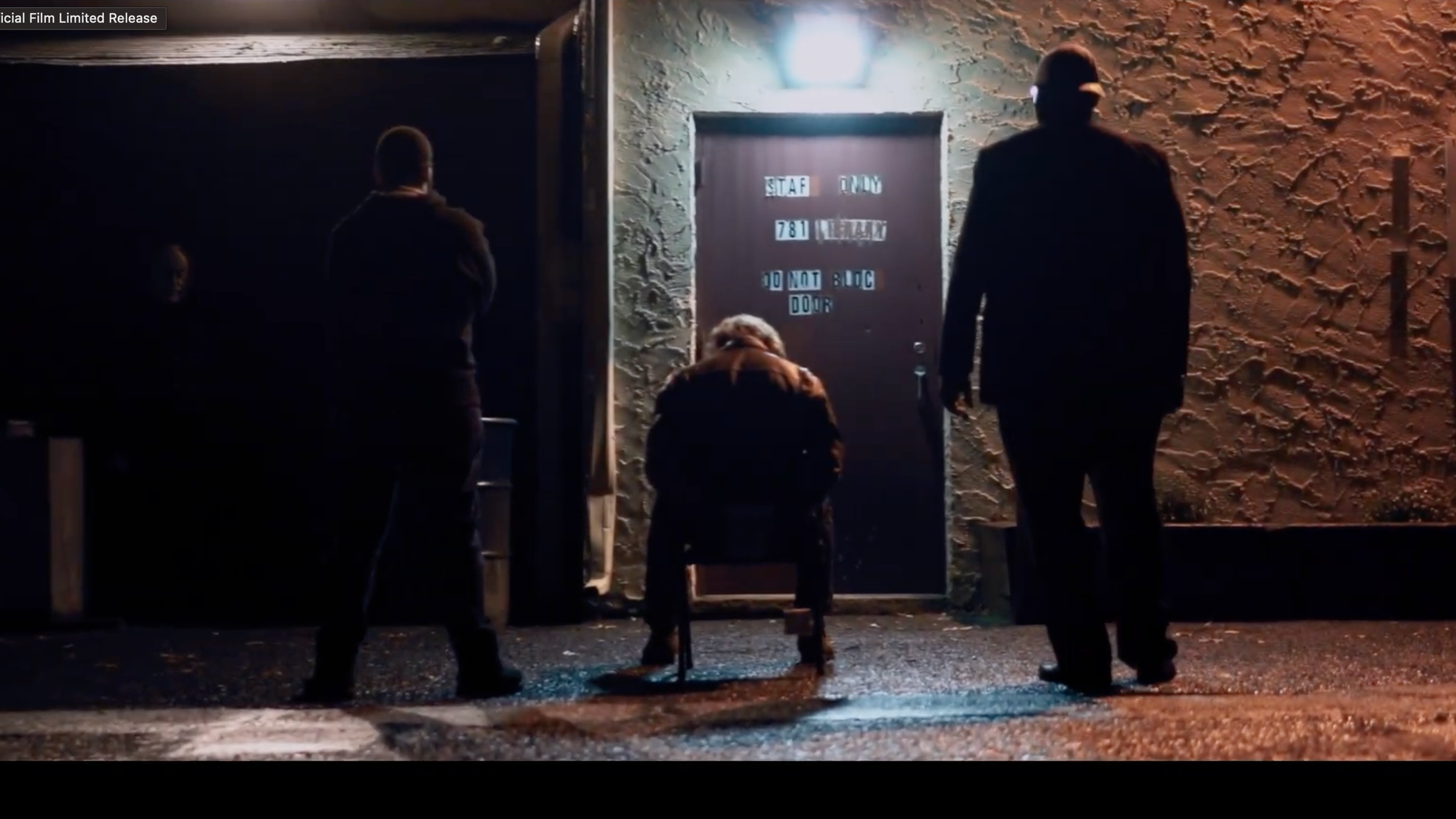 Four people are standing outside a door at night. The door has a sign with stylized letters, some of which are partially obscured. The scene is dimly lit, and the area has textured walls and a gravel ground.