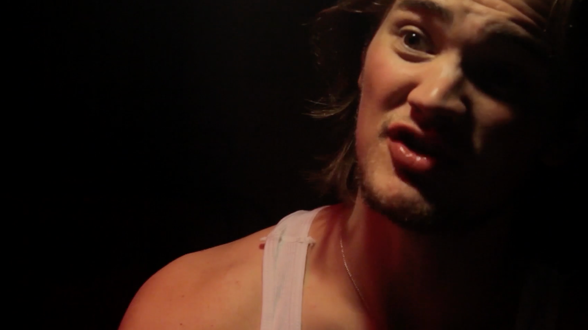 Close-up of a woman with light skin, shoulder-length wavy hair, wearing a sleeveless top, in a dimly lit setting.