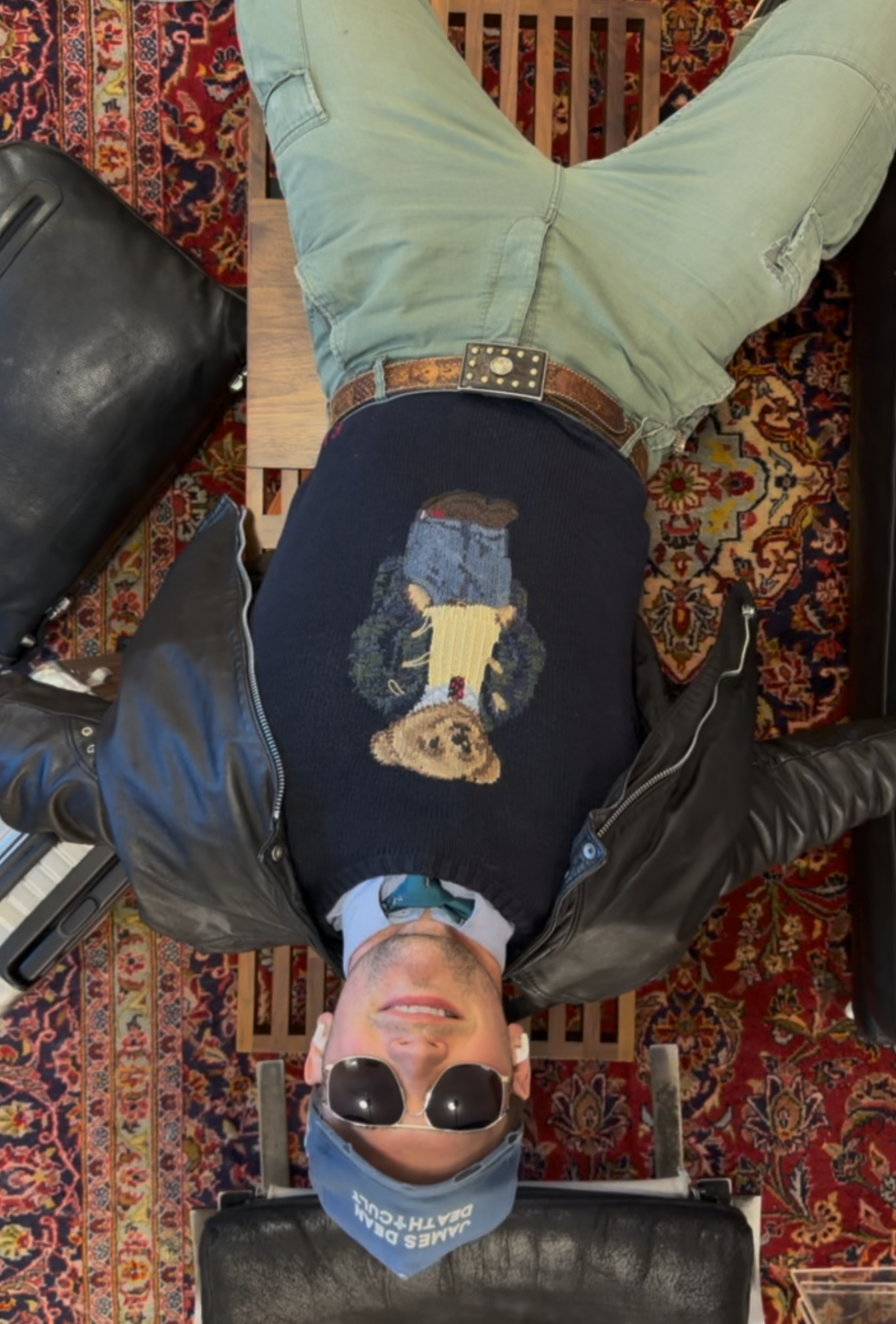 A person wearing sunglasses, a face mask, a blue head covering, a black jacket, and a graphic shirt with a wolf design, lying on a massage table with a patterned rug underneath, a black leather bag beside, and metal bars at the foot of the table.