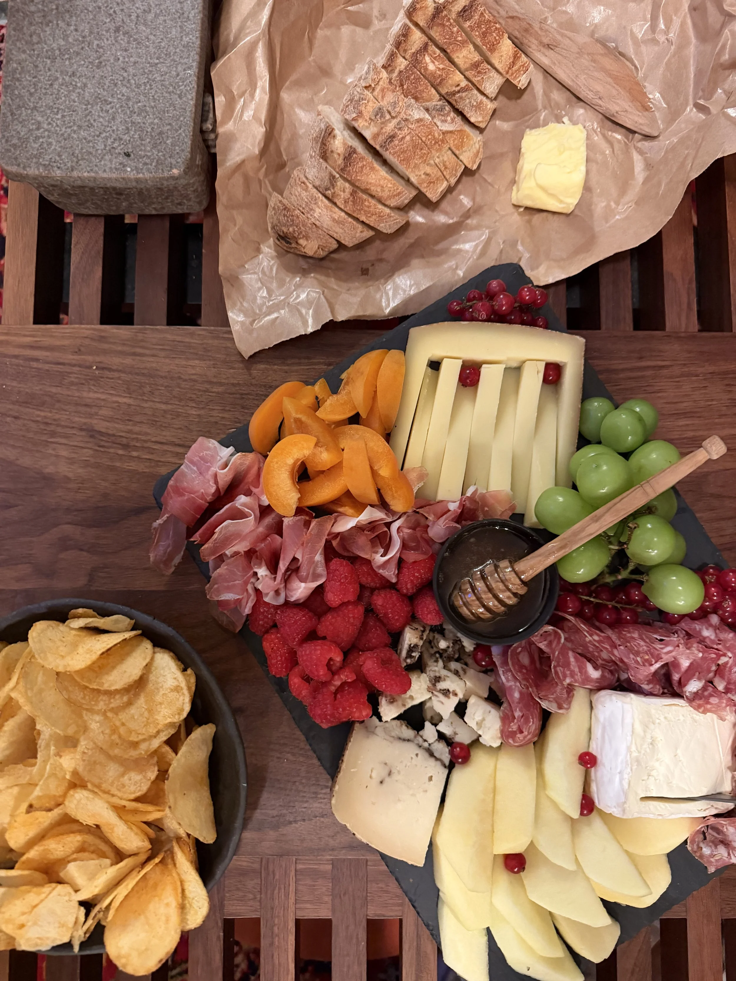 An assortment of sliced bread, cheeses, fresh fruits including grapes and raspberries, cured meats, honey, and potato chips on a wooden table.