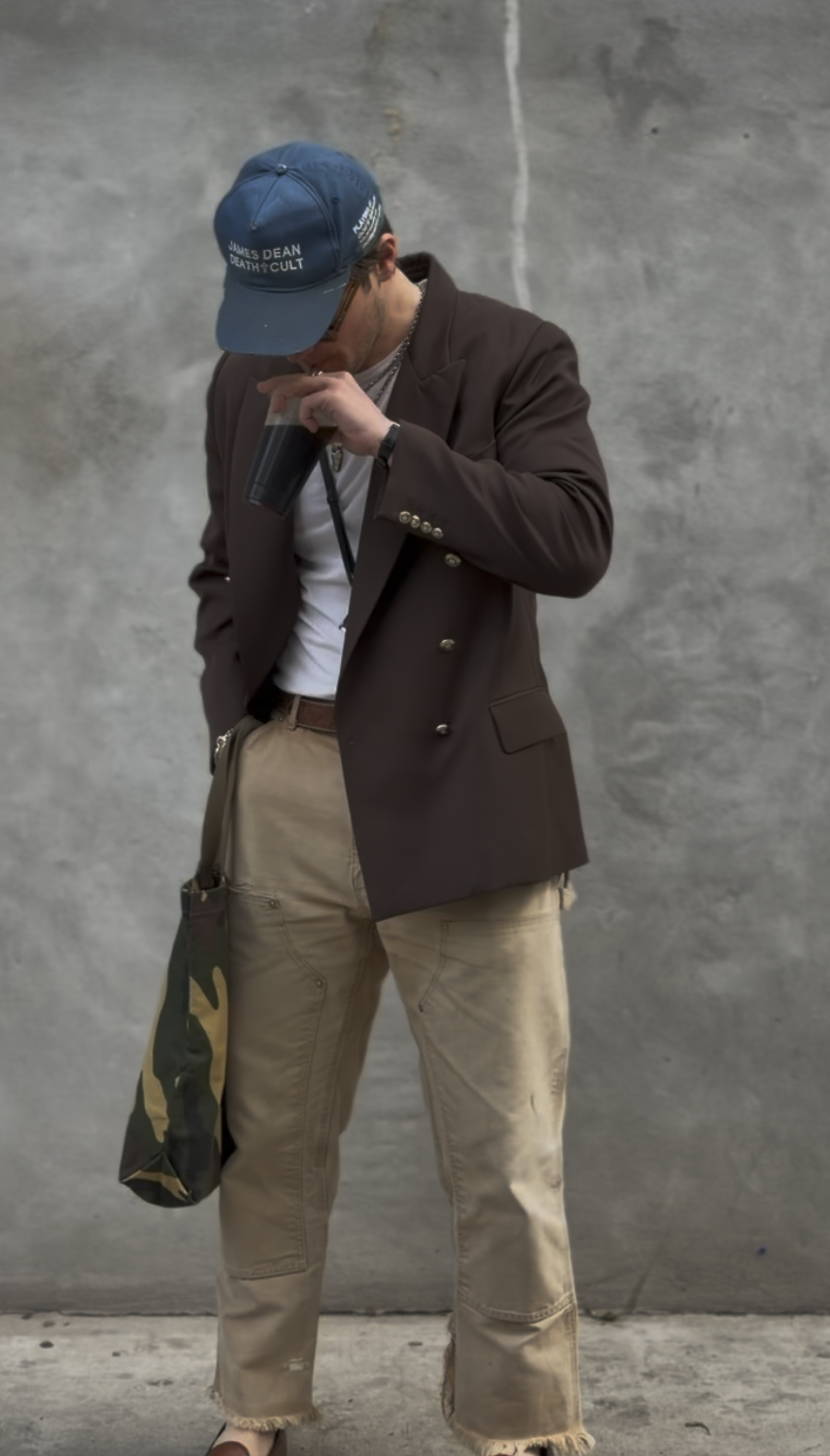 A person standing against a plain, gray concrete wall, wearing a dark brown blazer, beige pants, a blue cap, and glasses, holding a black cup to their mouth.