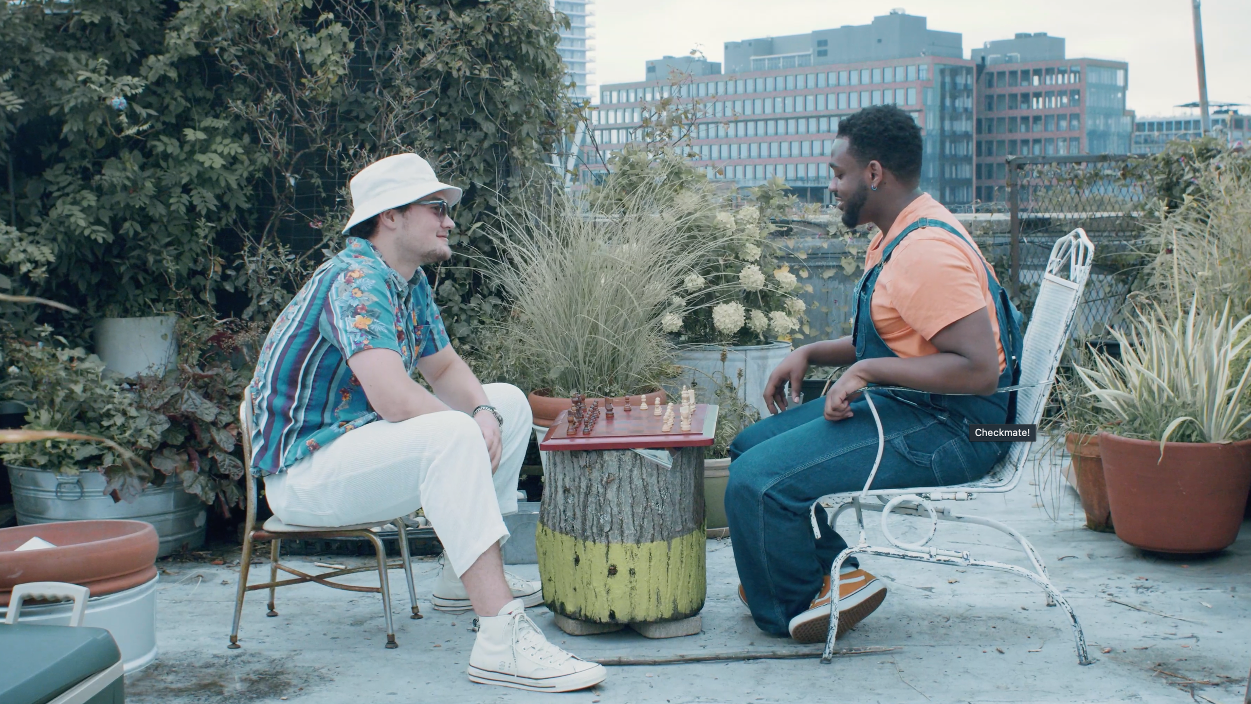 Two men playing chess outdoors on a rooftop garden with potted plants and tall buildings in the background.