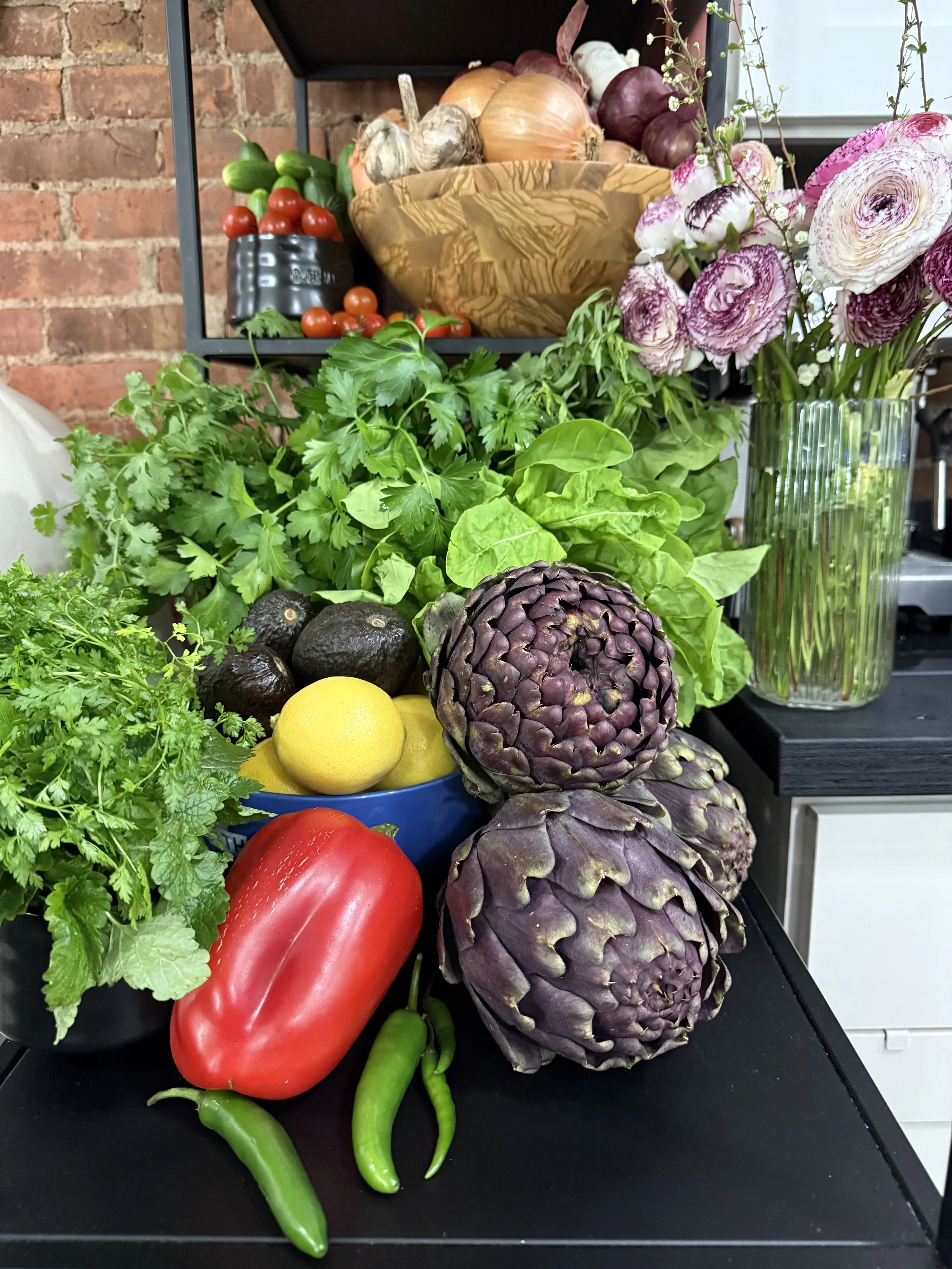 Arrangement of fresh vegetables including cilantro, a red bell pepper, green chili peppers, an avocado, lemons, artichokes, tomatoes, onions, and a bouquet of pink and white ranunculus flowers in a glass vase, with a background of brick wall and blac