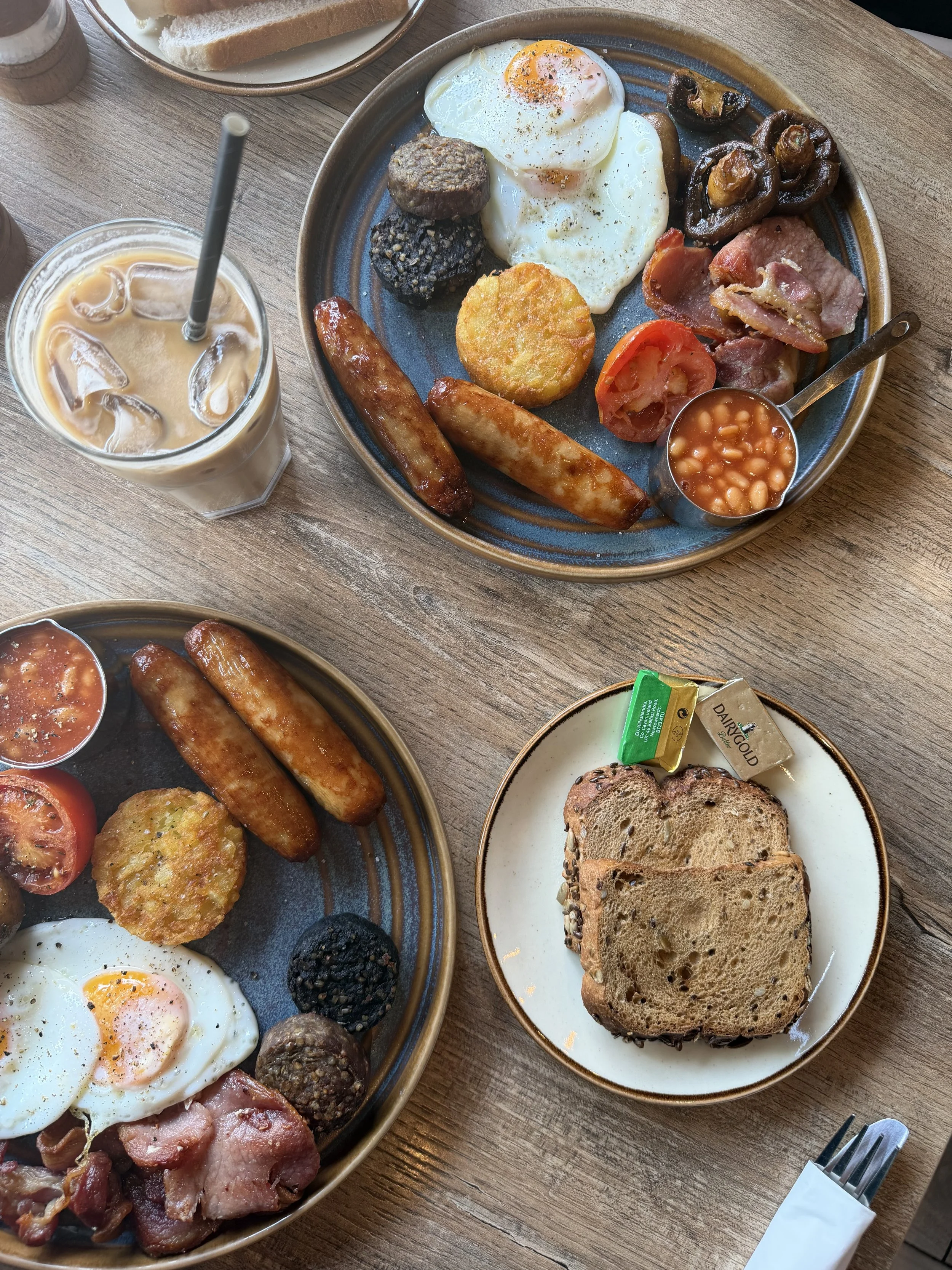 A breakfast plate with two fried eggs, sausages, bacon, hash brown, mushrooms, tomato, black pudding, and baked beans. A glass of iced coffee and a side of toast are also visible.