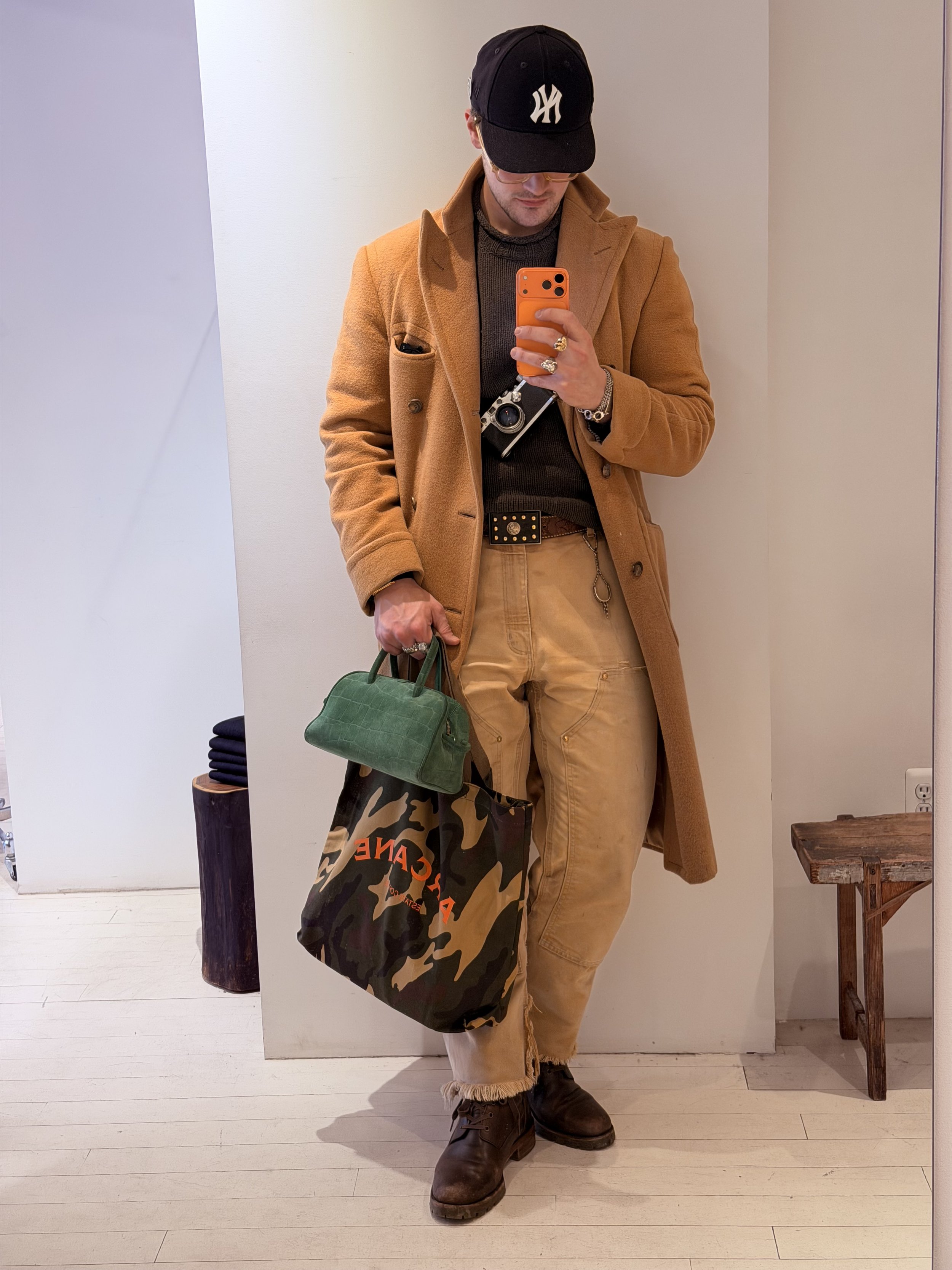 Person wearing a black New York Yankees cap, tan coat, gray sweater, tan pants, and brown boots, holding a green bag and a shopping bag, taking a mirror selfie in a minimalistic room.