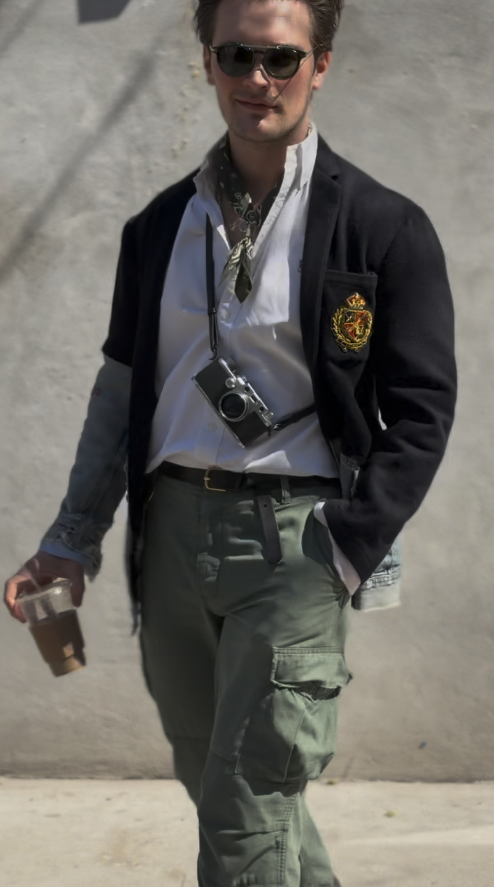 A man wearing sunglasses, a black jacket with an emblem on the sleeve, a white shirt, and cargo pants walks outside holding a coffee cup in one hand and a camera around his neck.