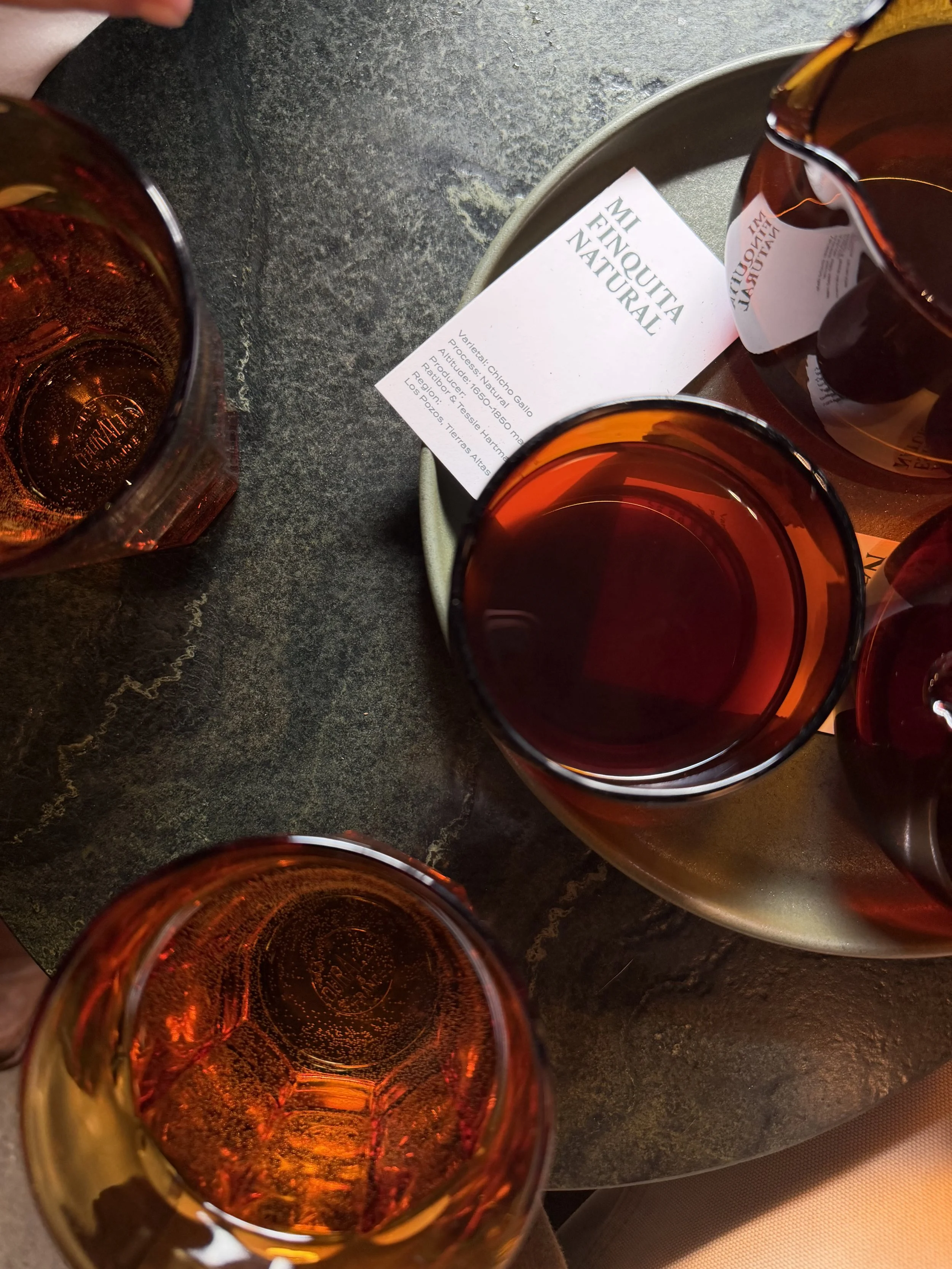 Four glasses of amber-colored liquid on a dark table, with a white informational card labeled 'Mi Menquita Natural' in the background.