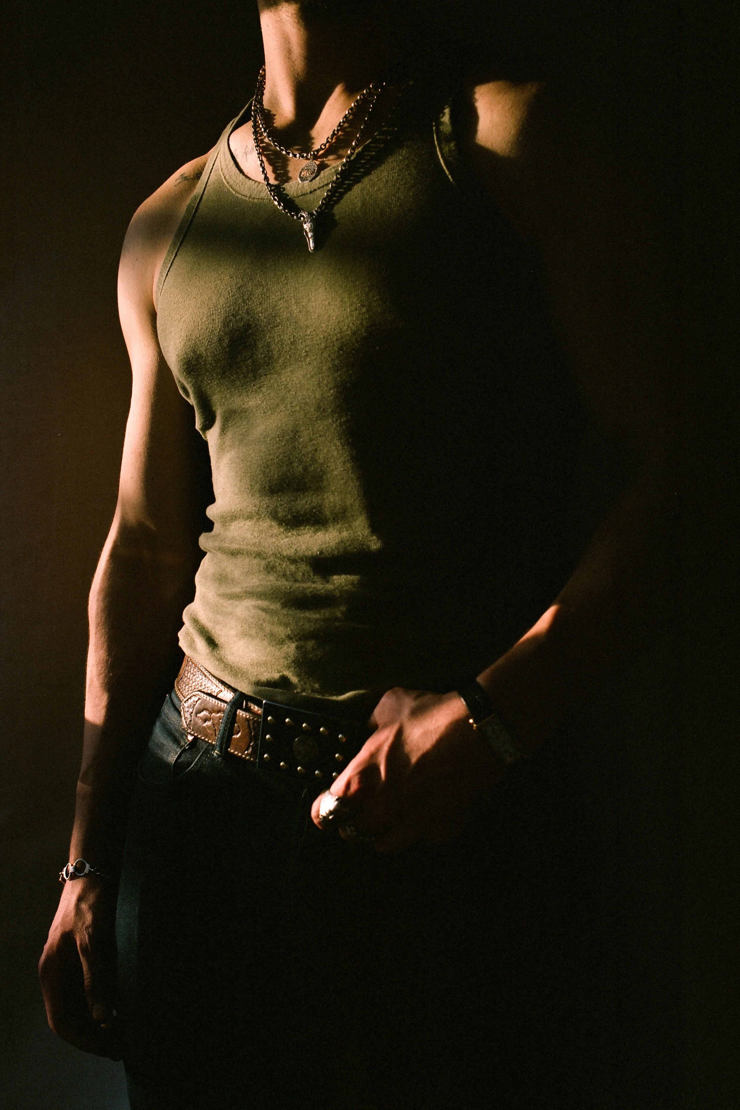 Part of a person wearing a green sleeveless shirt, multiple layered necklaces, black belt with studs, and a ring on the right hand, set against a dark background.