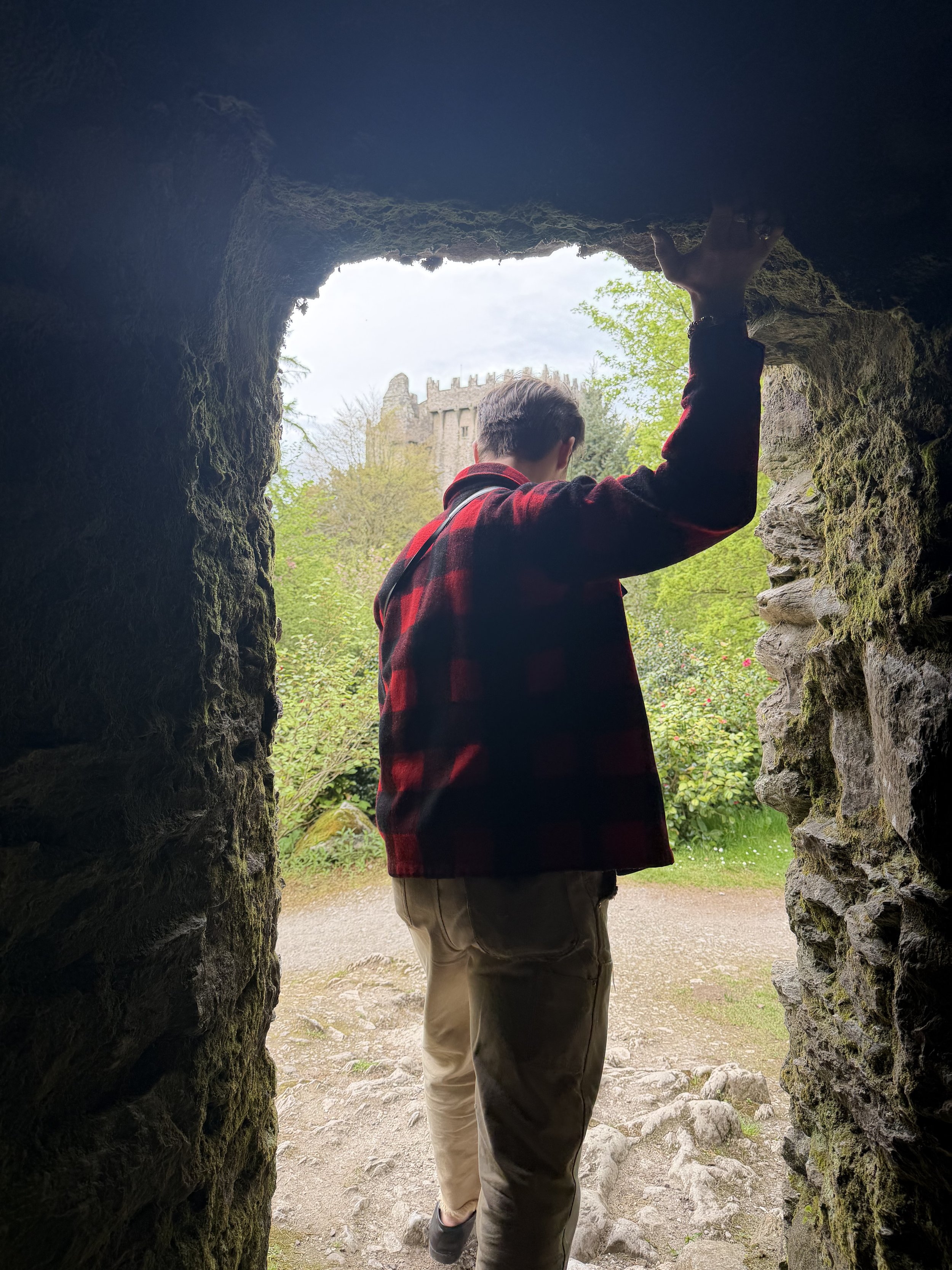 Person with short hair wearing a red and black plaid shirt and khaki pants, standing inside a stone tunnel, looking outside at a castle and trees, crossing a rocky path.