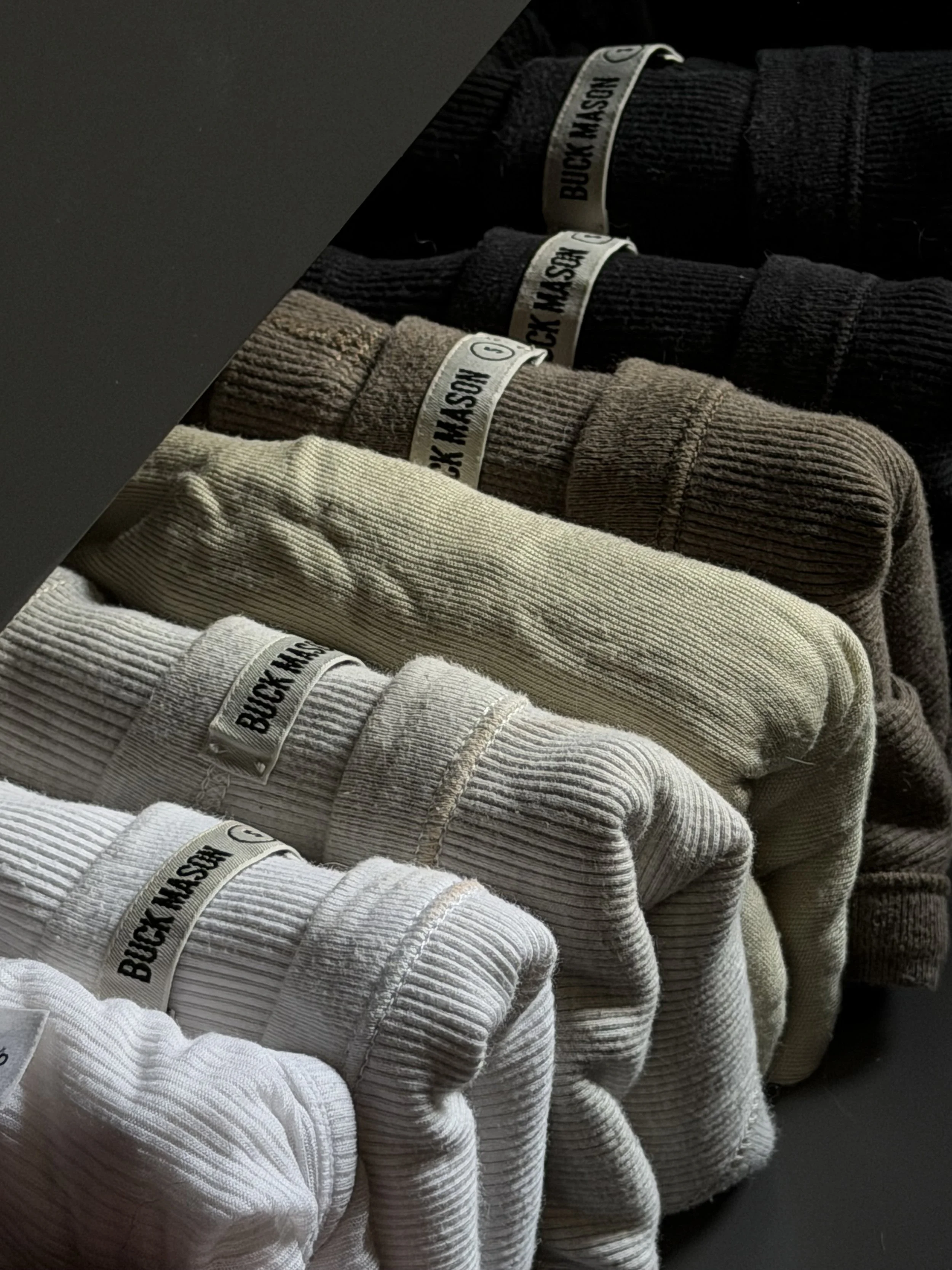 A display of four folded ribbed sweatshirts in black, beige, light gray, and white, each with a label reading 'BUCK MASON' attached to the collar.