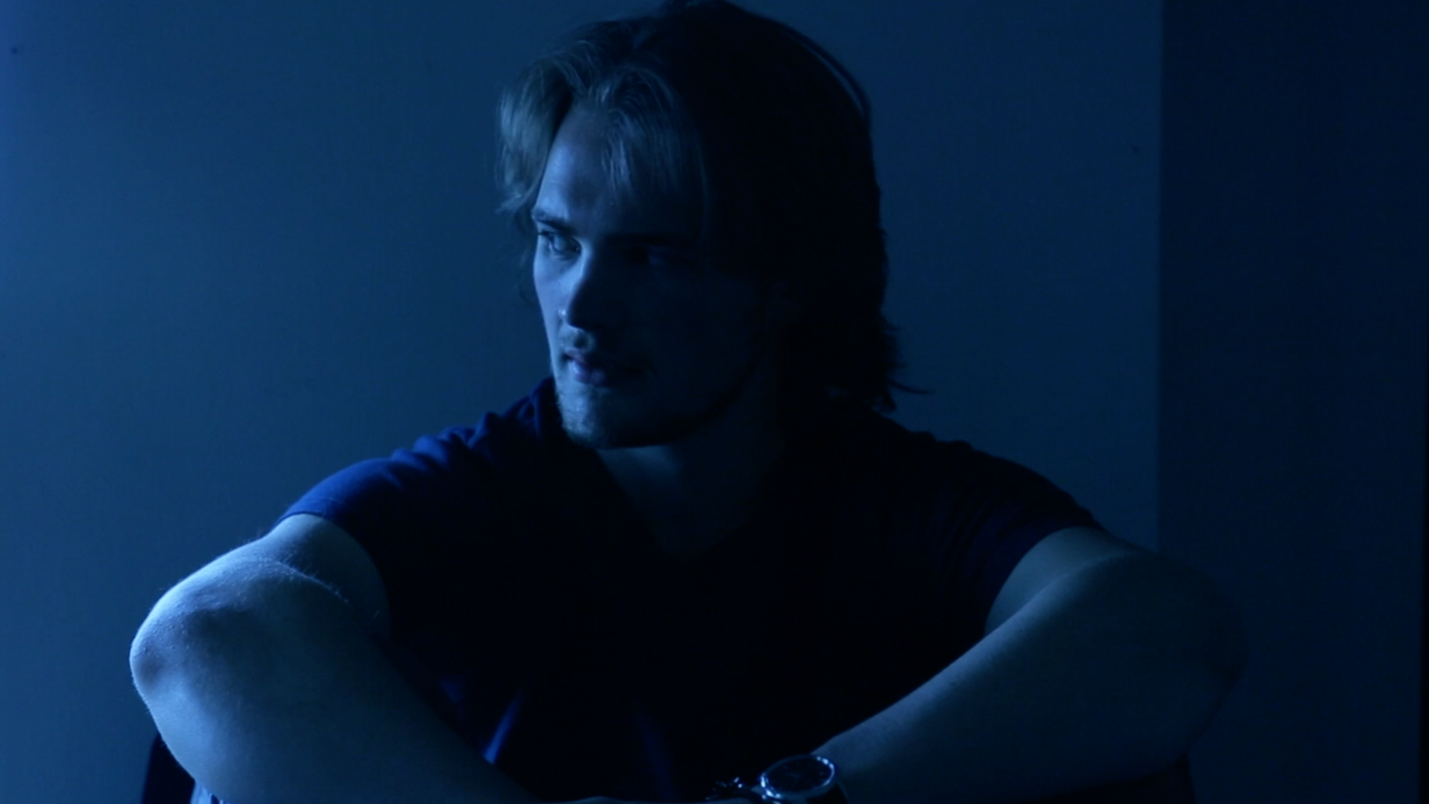A young man with long hair and a beard sitting in dim lighting, wearing a black t-shirt, with his arms crossed in front of him, looking to the side.