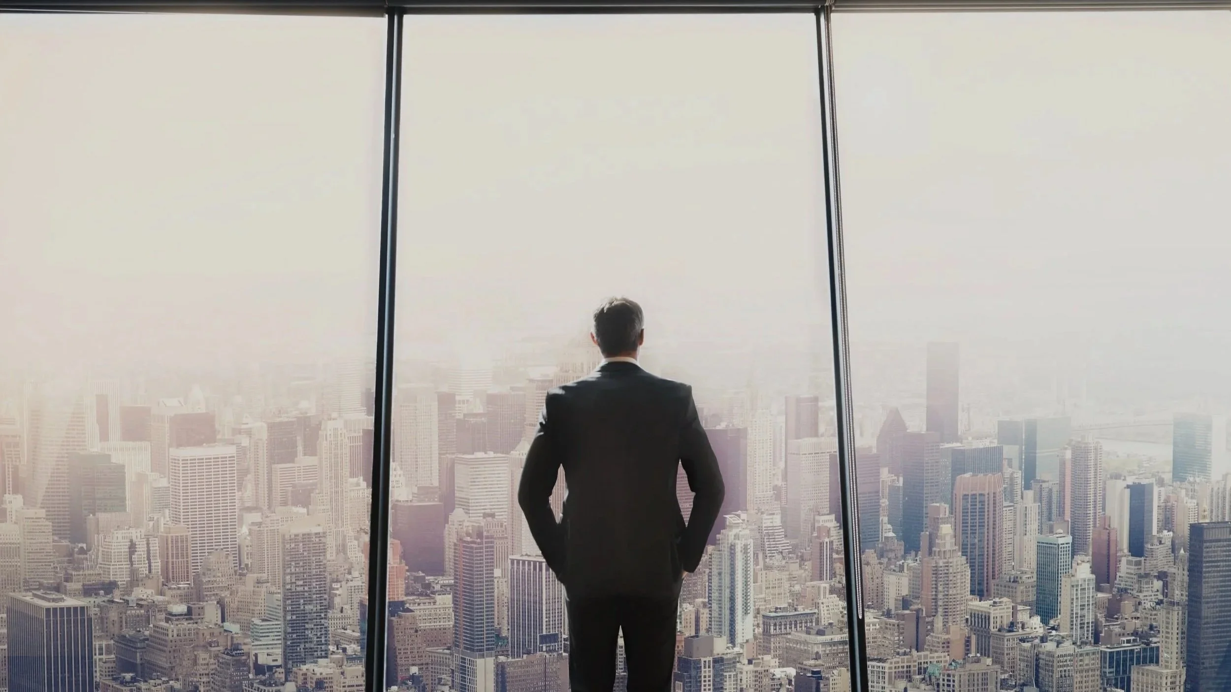 A man in a black suit stands with his back to the camera, looking out of a large window at a city skyline filled with skyscrapers.