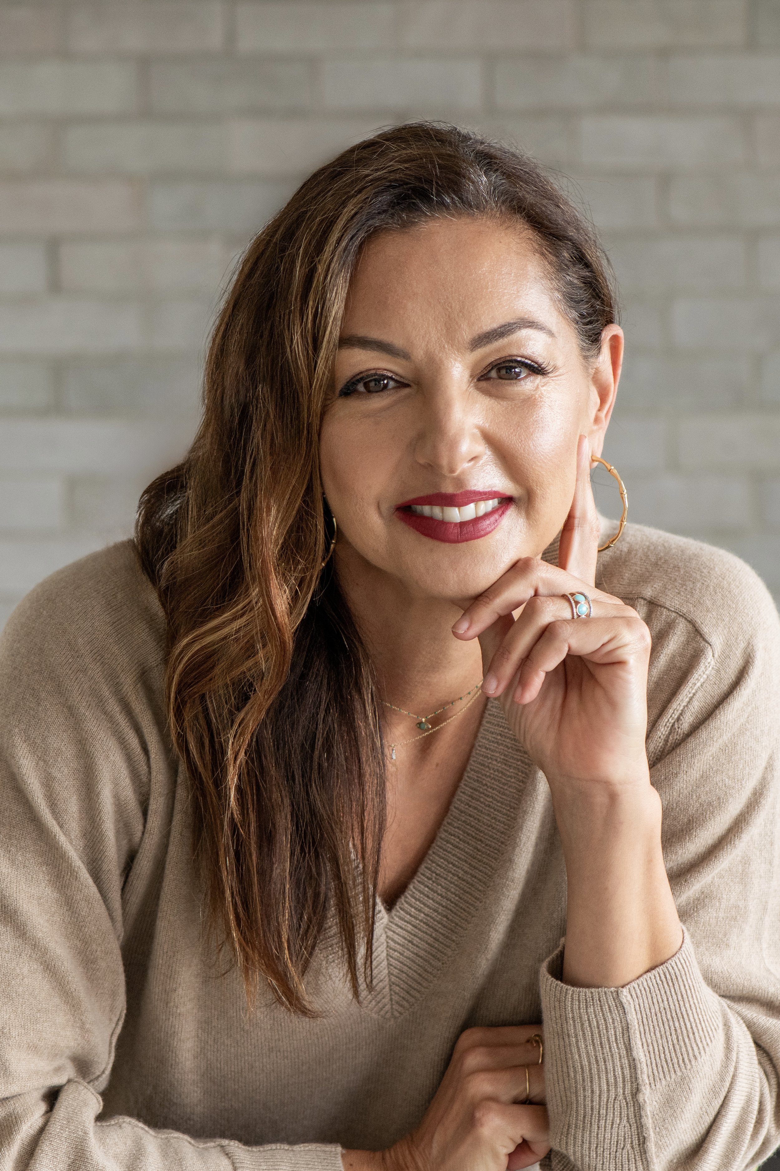 A woman with long brown hair, wearing a beige sweater, gold hoop earrings, and jewelry, smiling with her hand near her face against a light brick wall background.