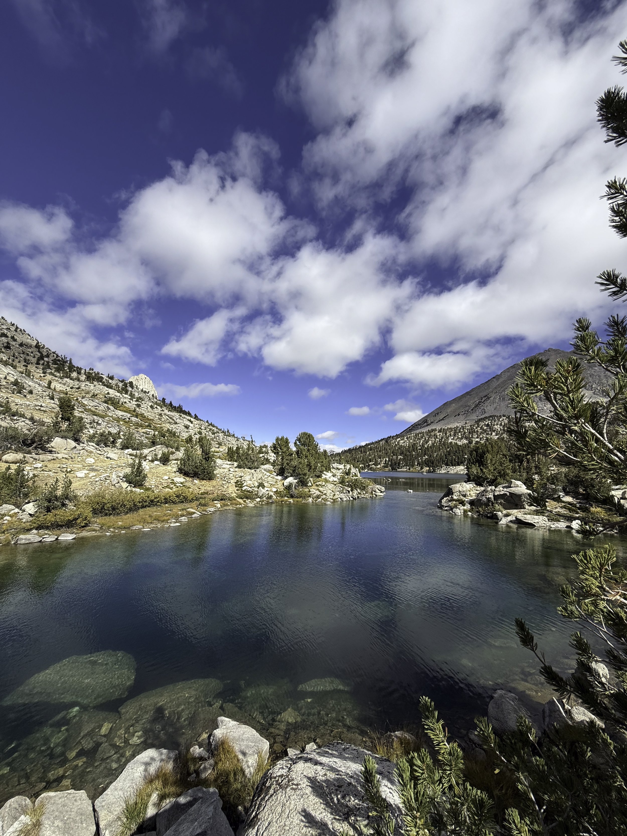 Scenic view of a clear mountain lake with rocks and trees along the shoreline, hills with sparse vegetation, and a partly cloudy sky overhead.