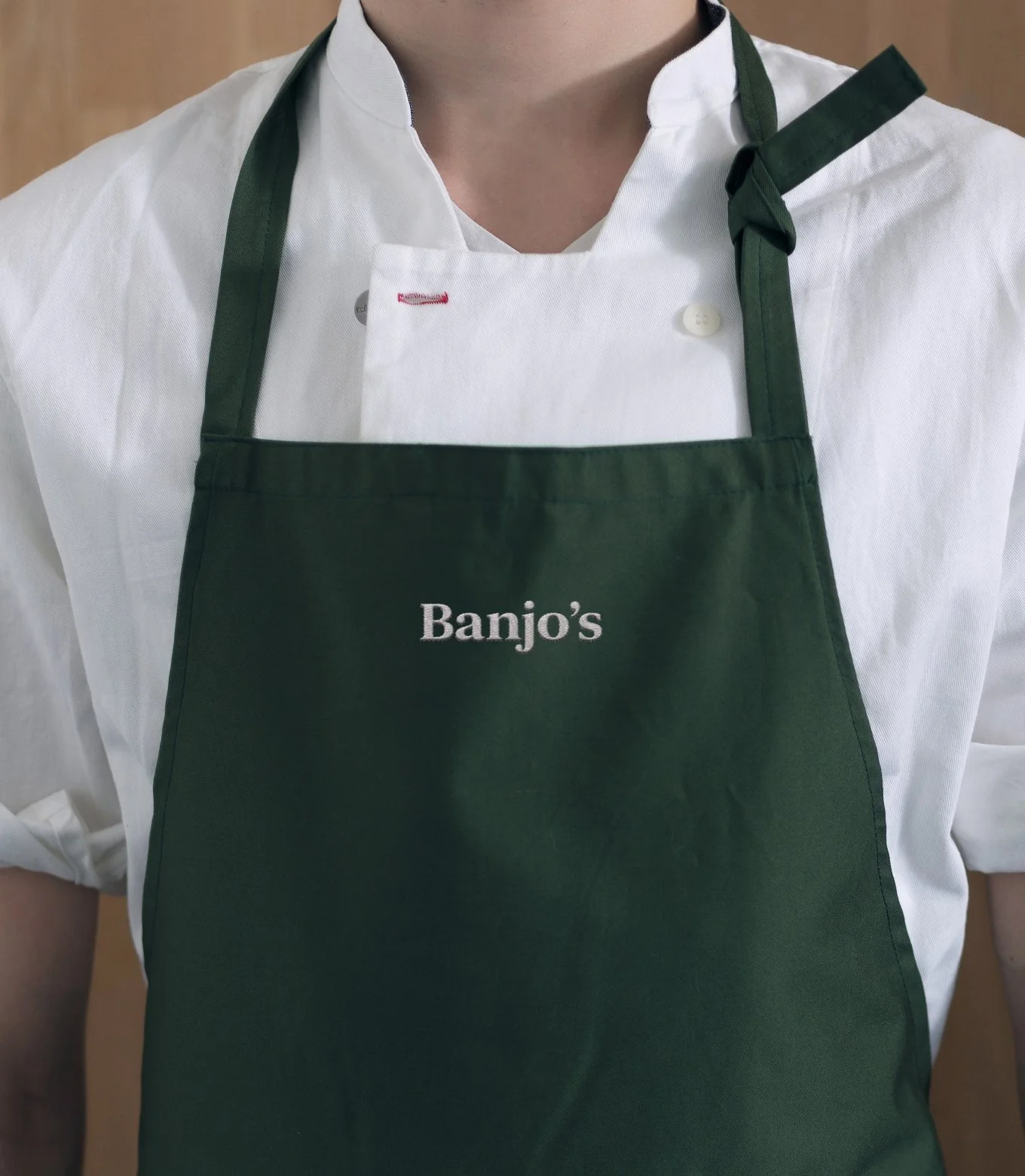Close-up of a person wearing a white chef's coat and a green apron with the word 'Banjo's' embroidered on it.