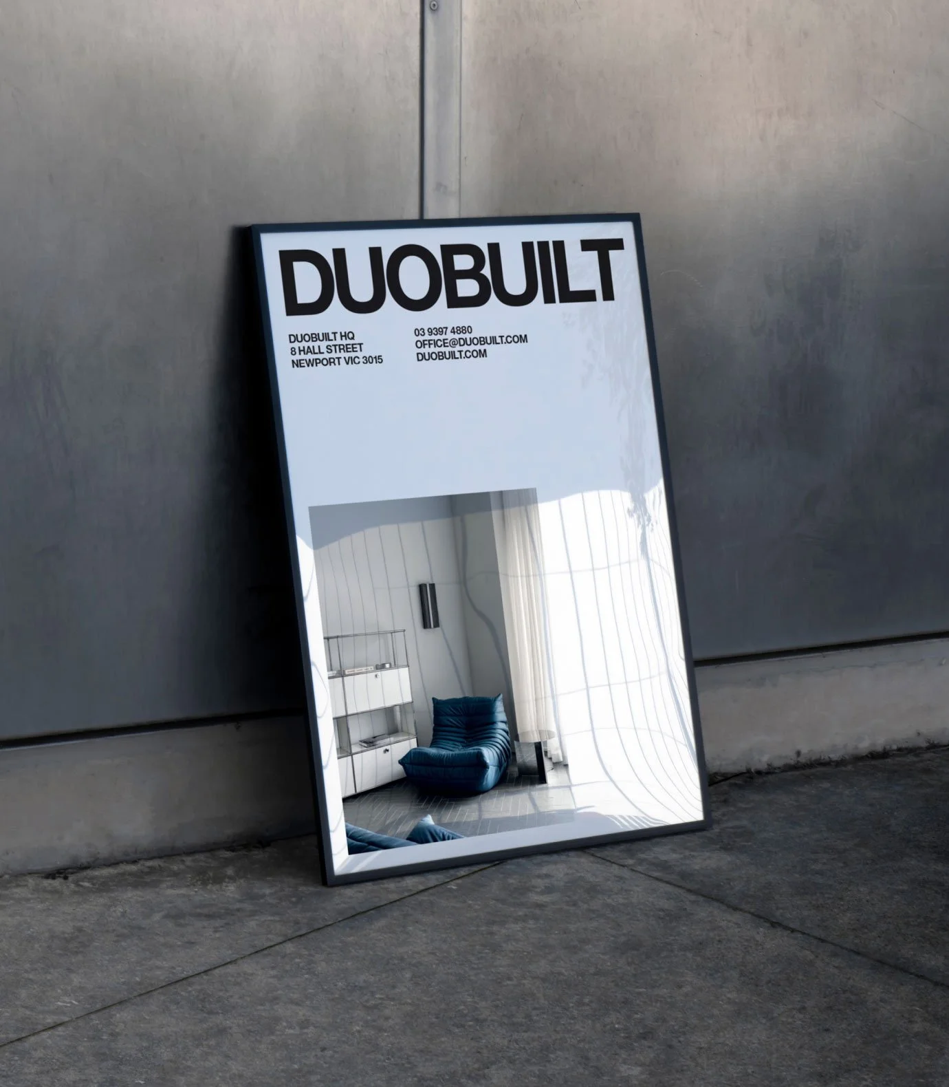 A large mirror with a sign on top reading DUOBUILT, placed on a concrete floor against a gray metal wall. The reflection shows a room with a blue chair, a white cabinet, large windows, and white curtains.