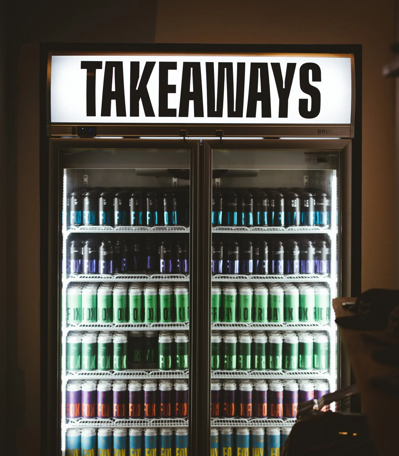 A refrigerator with a large illuminated sign above it that says "TAKEAWAYS." Inside the fridge, there are rows of cans with colorful labels in blue, green, purple, orange, and yellow.