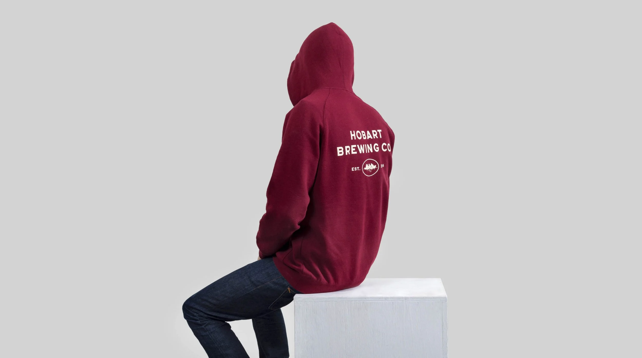 Person sitting on a white block, wearing a red hoodie with 'HOBART BREWING CO' printed on the back, and dark jeans, facing away from the camera, against a light gray background.