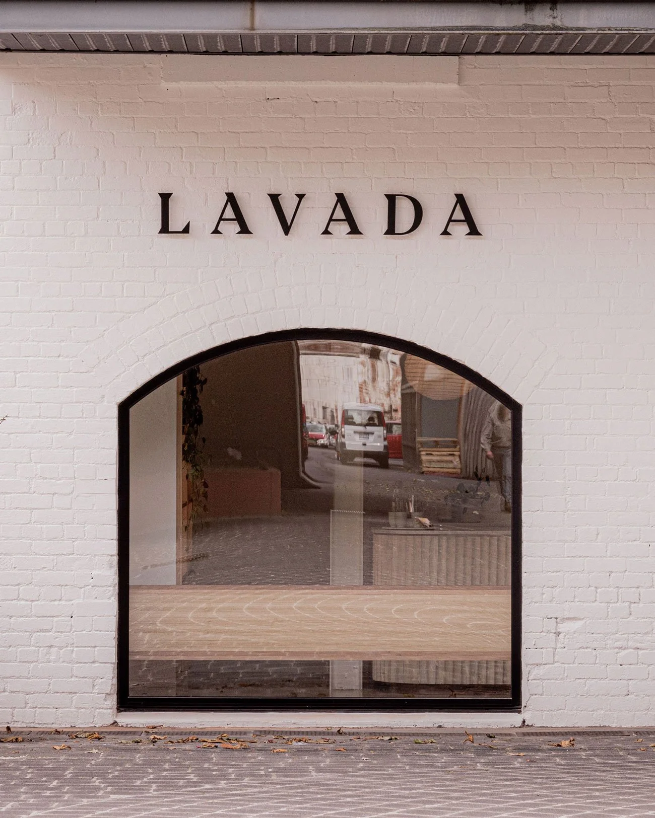 White brick building with a large arc window and the word 'LAVADA' on the wall above it. Reflections of a street scene with cars and people can be seen in the window.