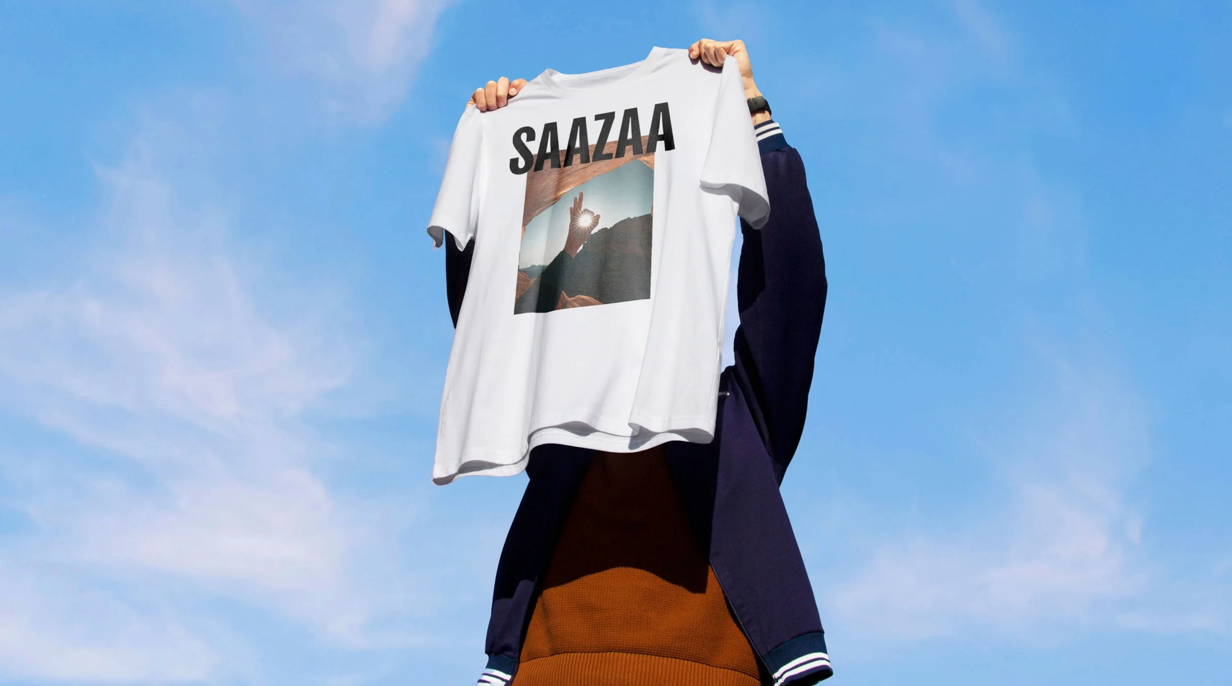 Person holding a white T-shirt with a graphic of a hand making a heart gesture and the word 'SAAZAA' printed above, against a blue sky background.