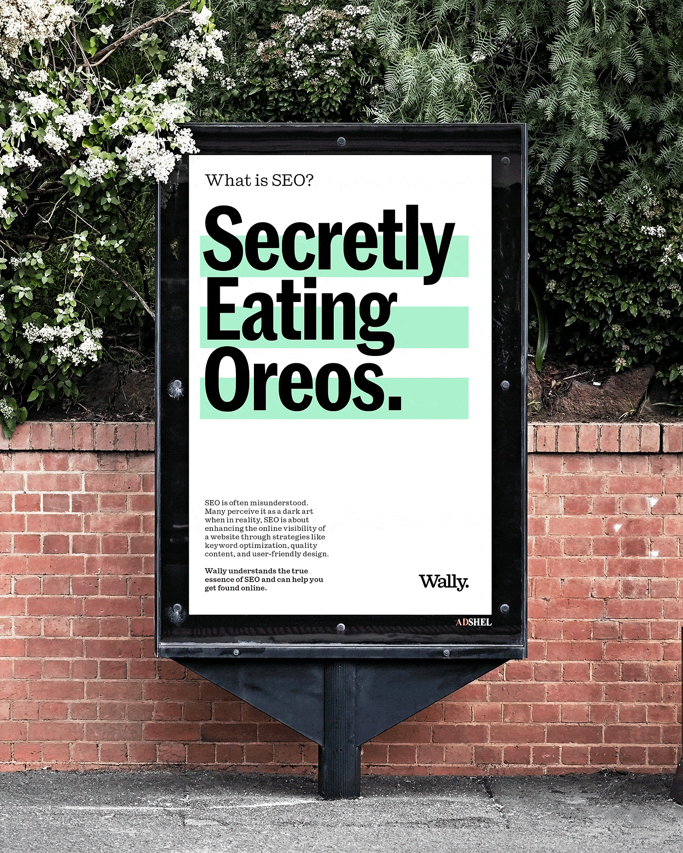 An outdoor advertising billboard with a white background and black and green text on a brick wall. The text reads 'What is SEO? Secretly Eating Ores.' Small text below explains SEO and a signature that says 'Wally.' The billboard is to the right of white flowering bushes and green foliage.